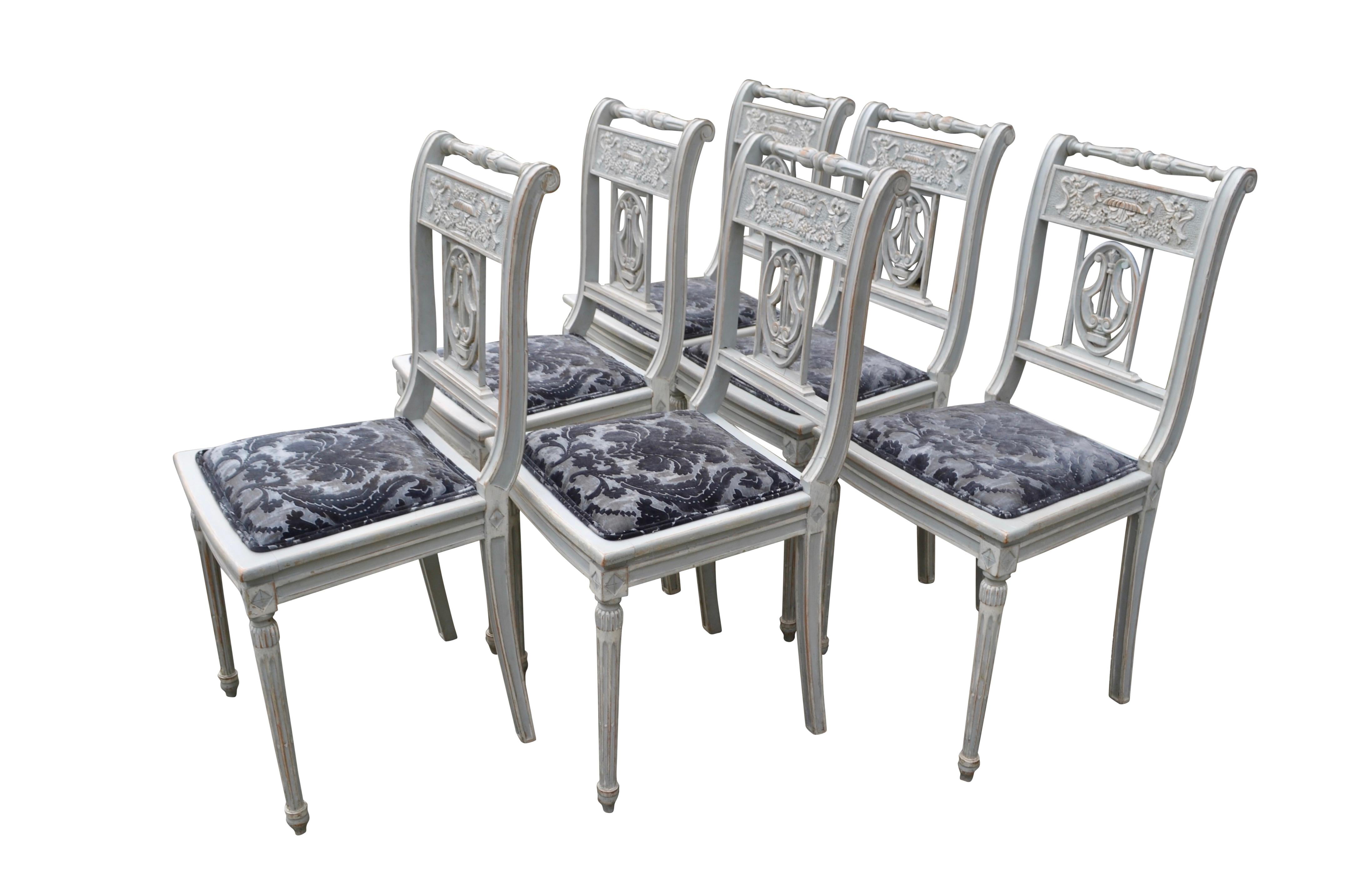 A very decorative set of six dining chairs of classical Directoire style, (referred to as Swedish Gustavian style). The open chair backs feature a central carved harp within an oval and above, a carved urn with flowers and garlands. The frames are