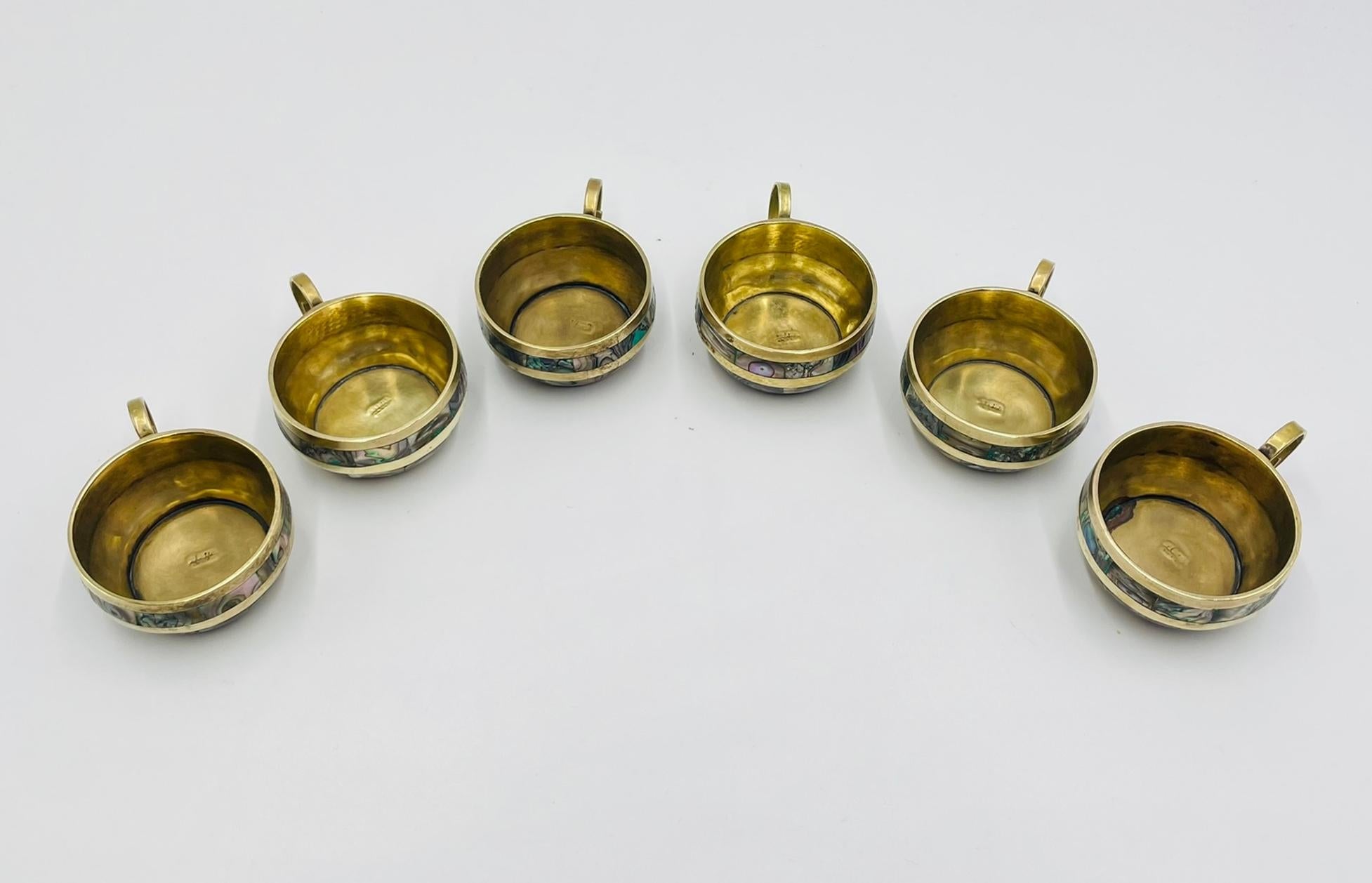 Elegante juego de seis tazas de té artesanales con platillos a juego, diseñadas y fabricadas en México durante la década de 1950. Cada pieza está bellamente fabricada en latón macizo con incrustaciones de concha de abulón, mostrando el refinado arte