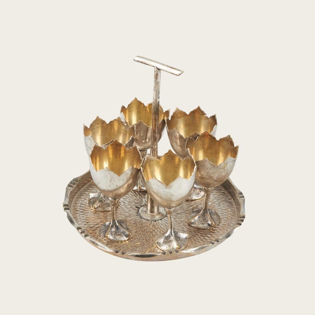 1950s, Sweden. This set includes six silver-plated brass egg cups with cracked egg-shell details and chicken foot bases, paired with a decorative tray featuring gauged accents. 