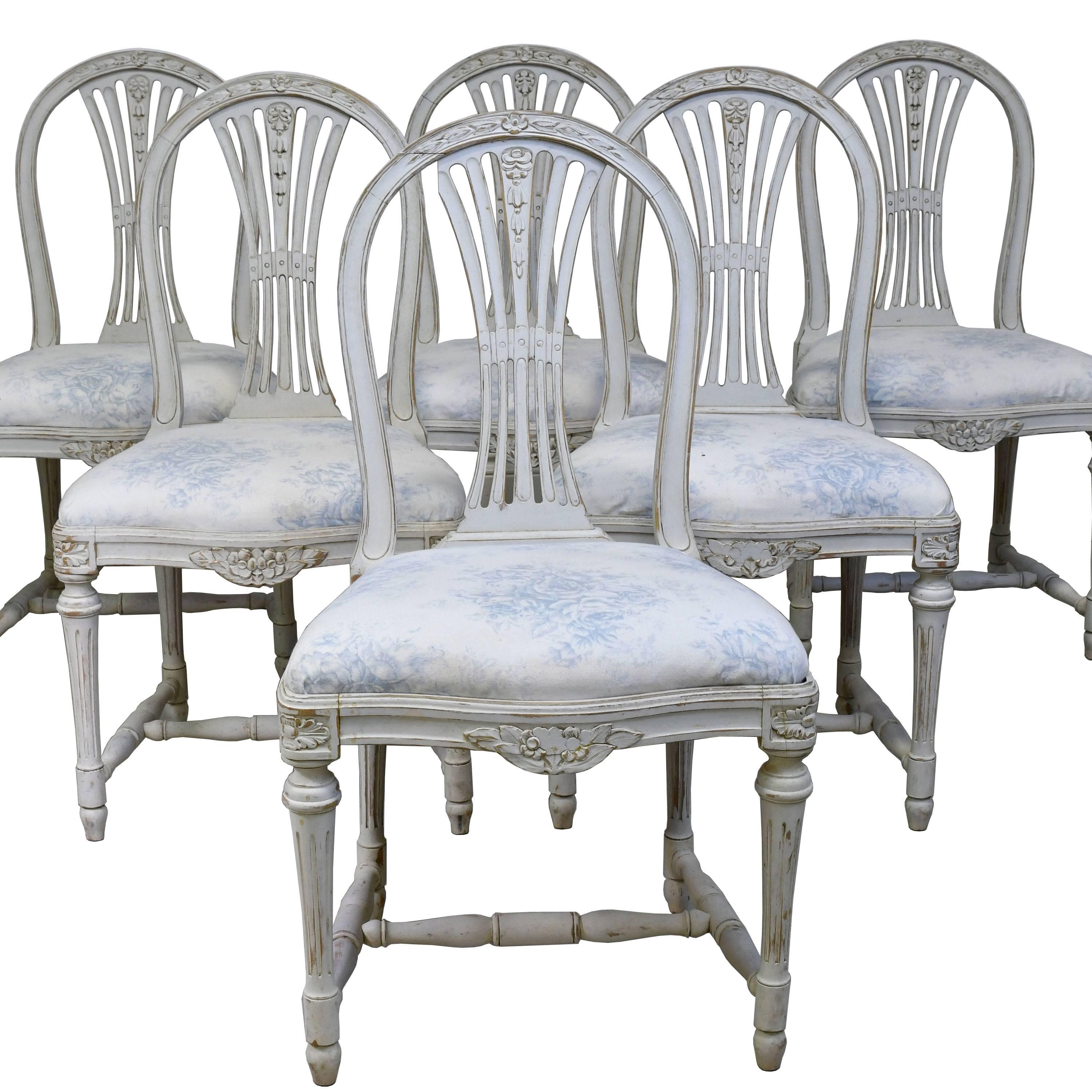 A lovely set of six (6) Gustavian-style dining chairs from the late 1800's with 18th century Lindome Gustavian style frames in a painted grey/white finish and with upholstered slip seat. Chairs have a carved sheaf-back, serpentine seat with foliate
