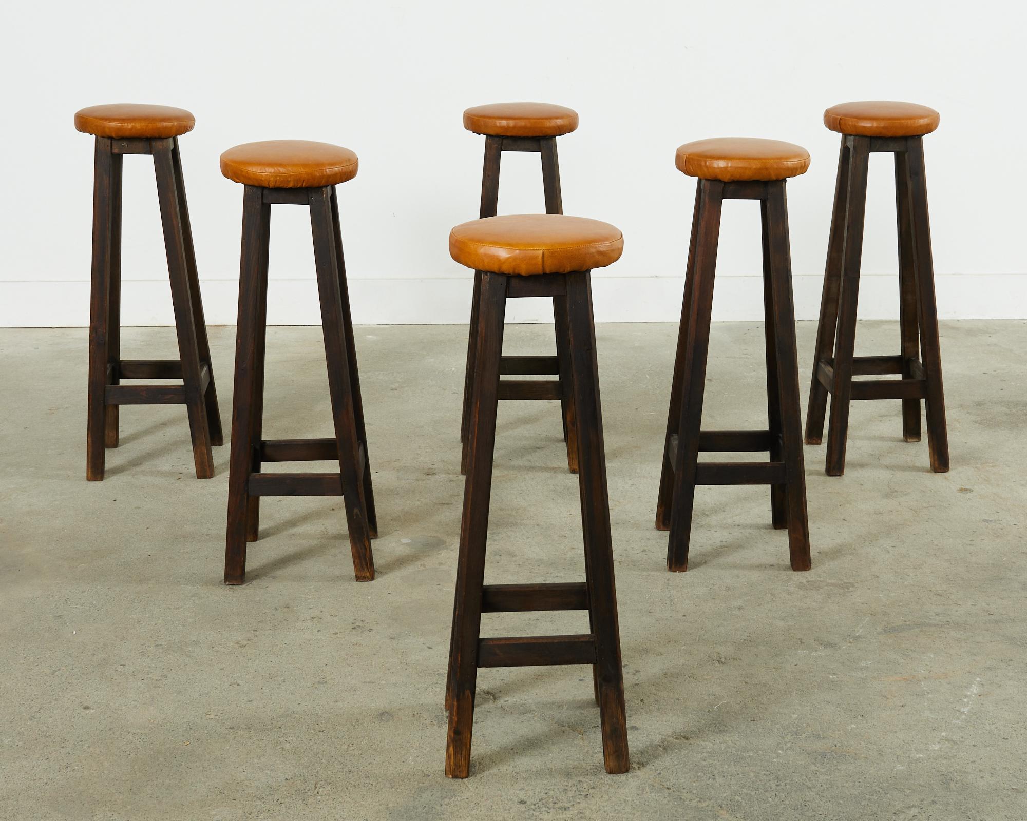 Rustic set of six matching pub style barstools featuring tall, narrow frames, crafted from oak with chambered legs conjoined by stretchers. Dark finish on the oak has an aged, faded patina. Topped with newly upholstered thick leather hide covered