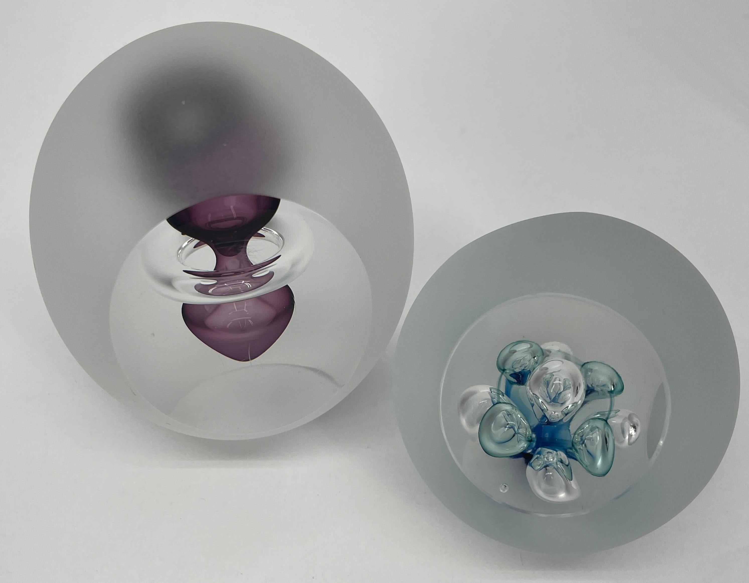 2 Modern Swedish Åhus glass paperweights. The signed paperweights are frosted glass with colorful interior and frosted glass surface. The larger piece is oval shaped with purple modern abstract floating glass interior. The smaller paperweight is