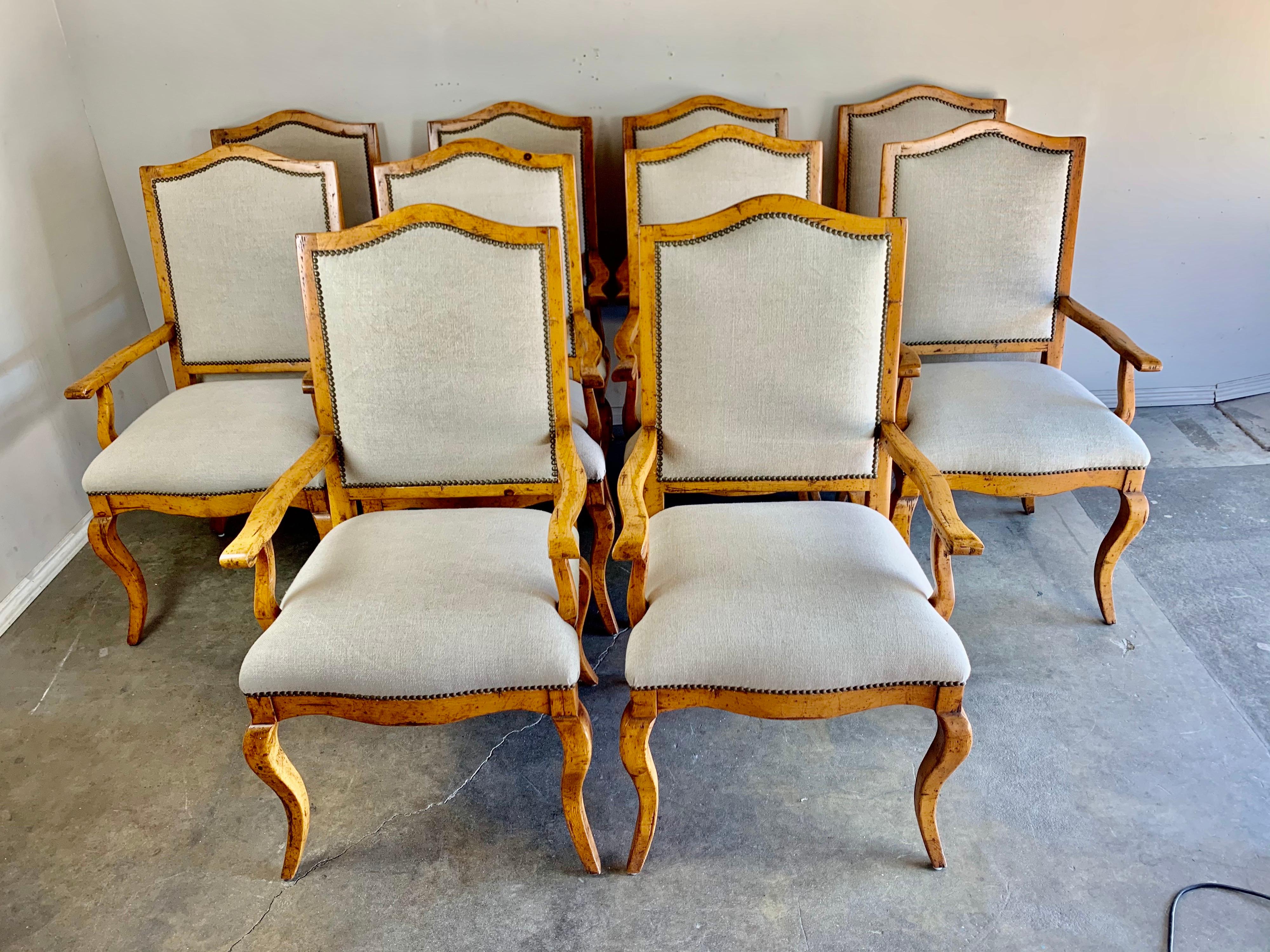 Set of ten pine dining armchairs that are newly upholstered in a washed Belgium linen with antique brass colored nailhead trim detail. The chairs have an eyebrow shaped top and stand on four cabriole shaped legs.