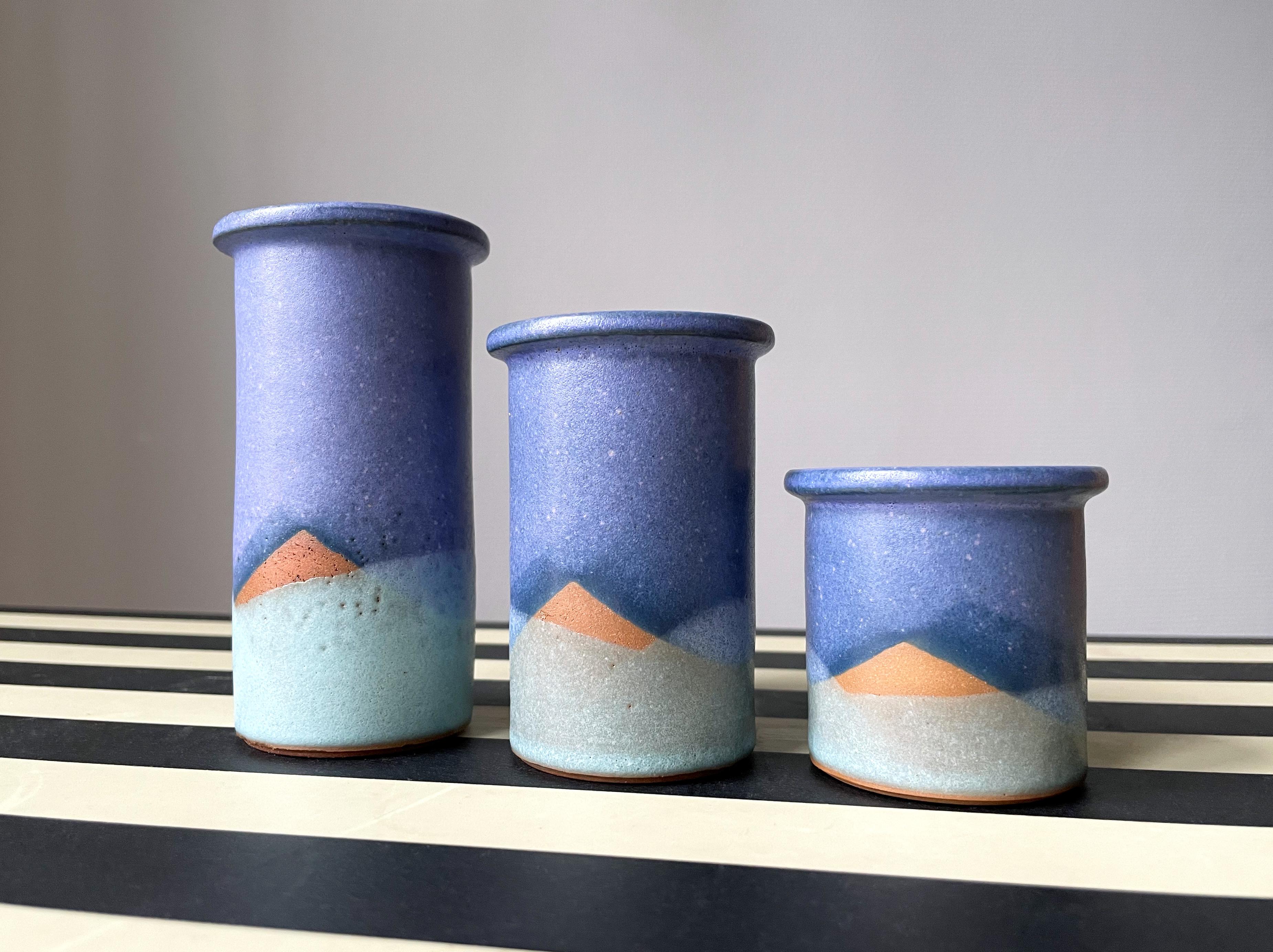 Set of three handmade Danish ceramic vases / candle sticks with graphic decor using the raw ceramic as part of a straight lined pattern resembling a landscape or a sunset. Same stylized decoration in blues and aqua green on all three vases. A small