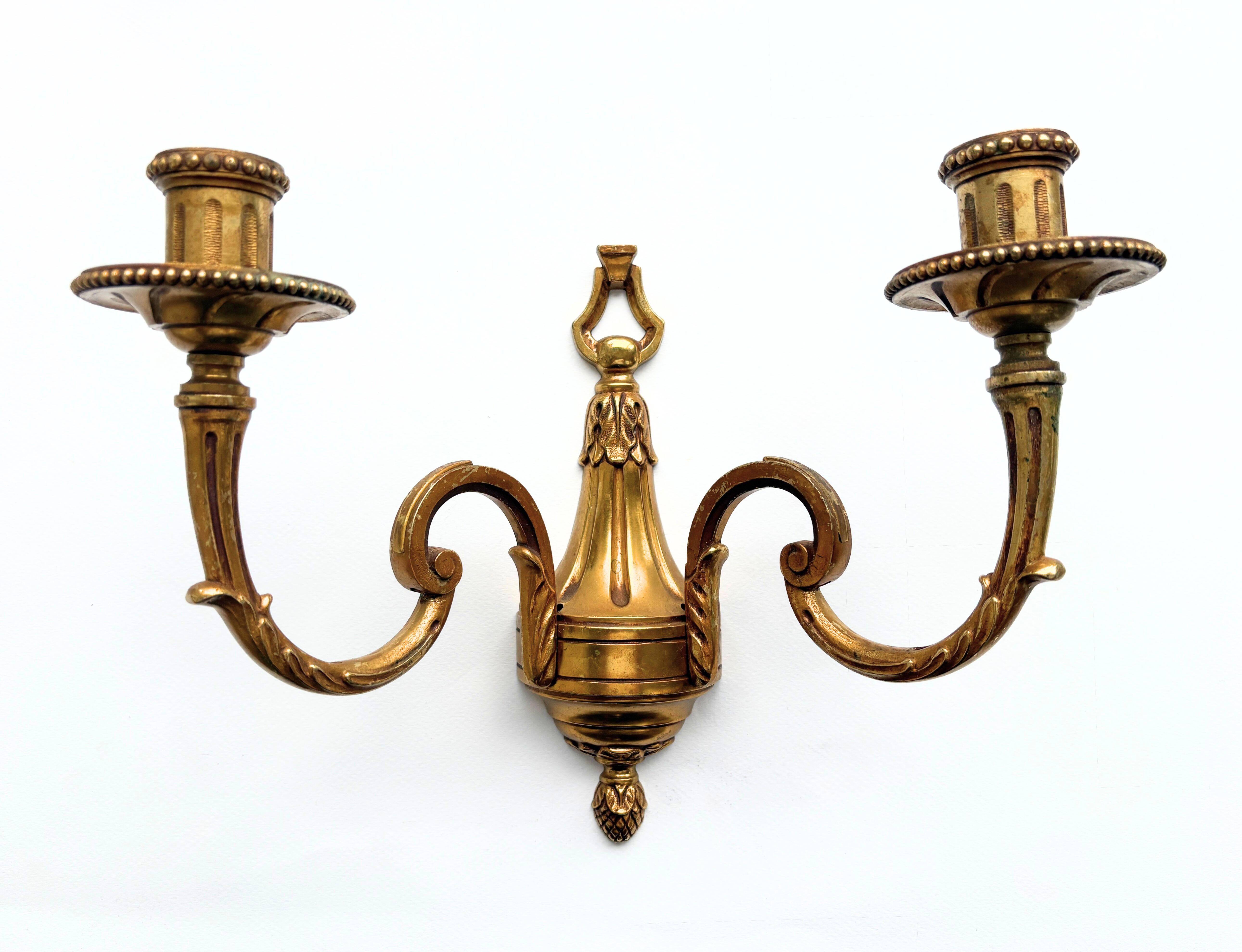 A set of three bronze wall sconces, made and sourced in France, dating from the 1960s—a period when Hollywood Regency interiors embraced the refined elegance of Neoclassical design. Each sconce features twin candle arms with graceful symmetry,