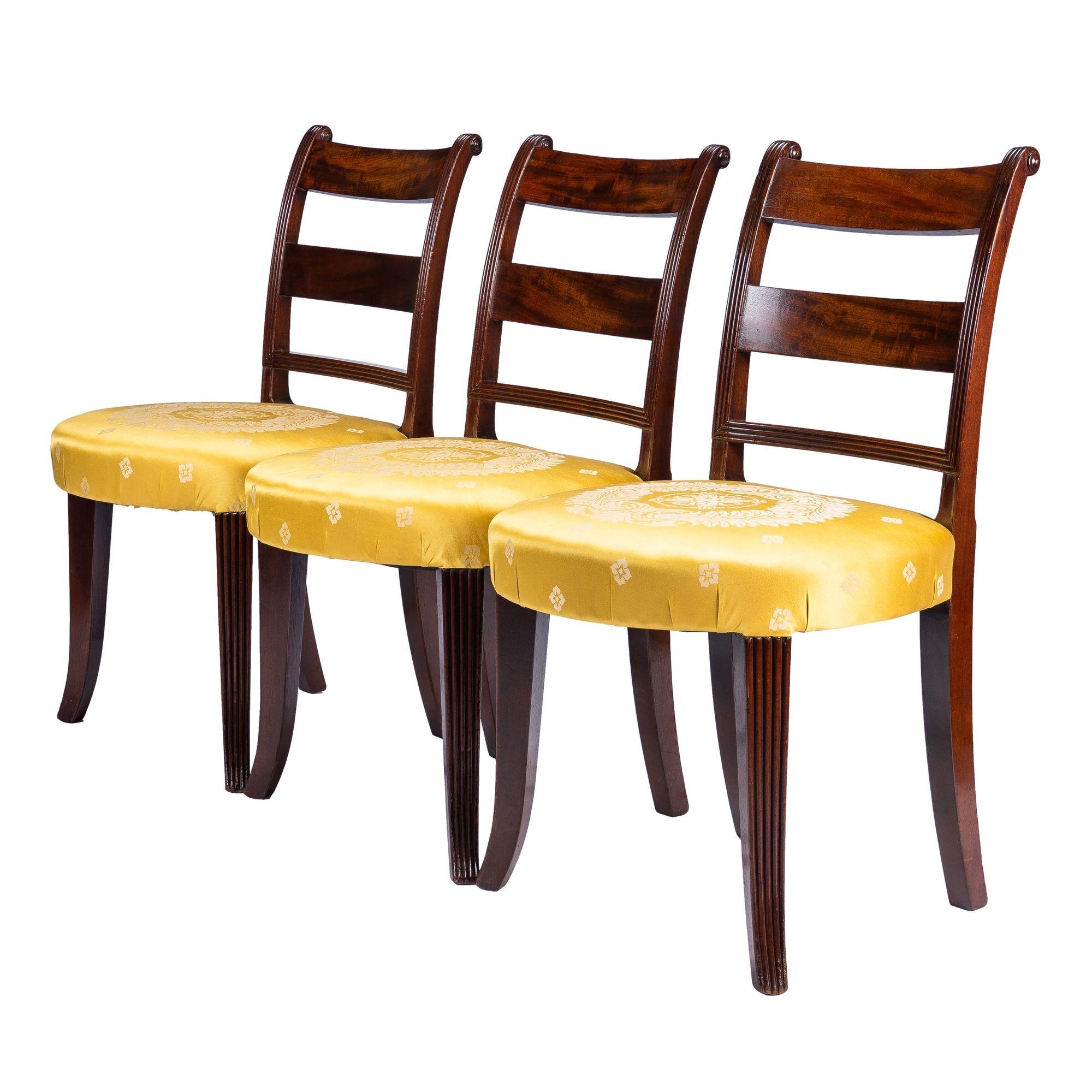 Set of three American mahogany upholstered seat side chairs on reeded sabre legs with flared feet. The London cabinetmakers book of this period describes a scroll back chair with a bell-shaped seat and sweeping Grecian front legs with a hollow below
