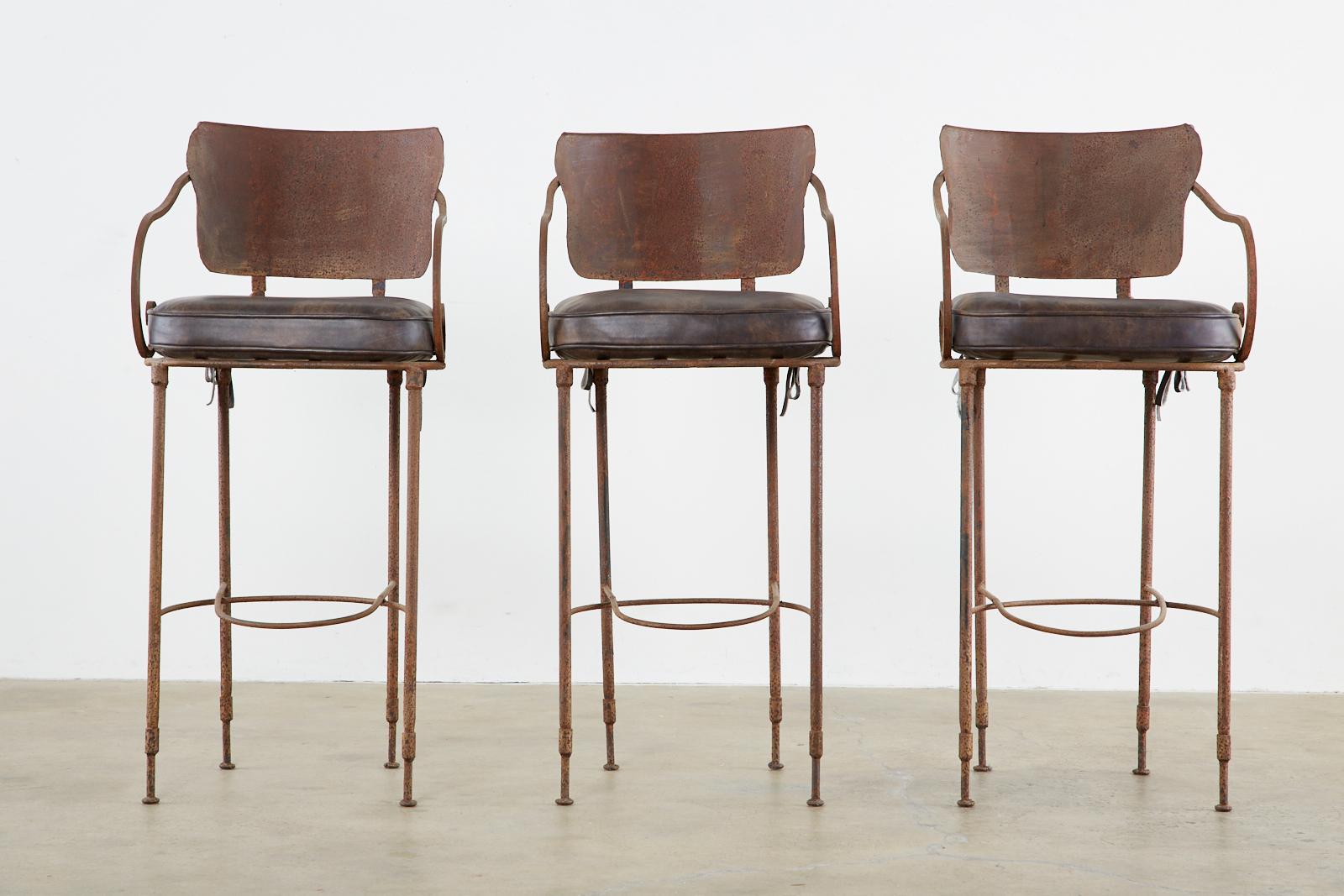 Rustic set of three iron and leather barstools hand forged by Arhaus. Each chair a masterpiece with a beautifully patinated iron finish. Topped with fitted leather seat cushions each stool is very heavy and sturdy with a unique handcrafted finish.