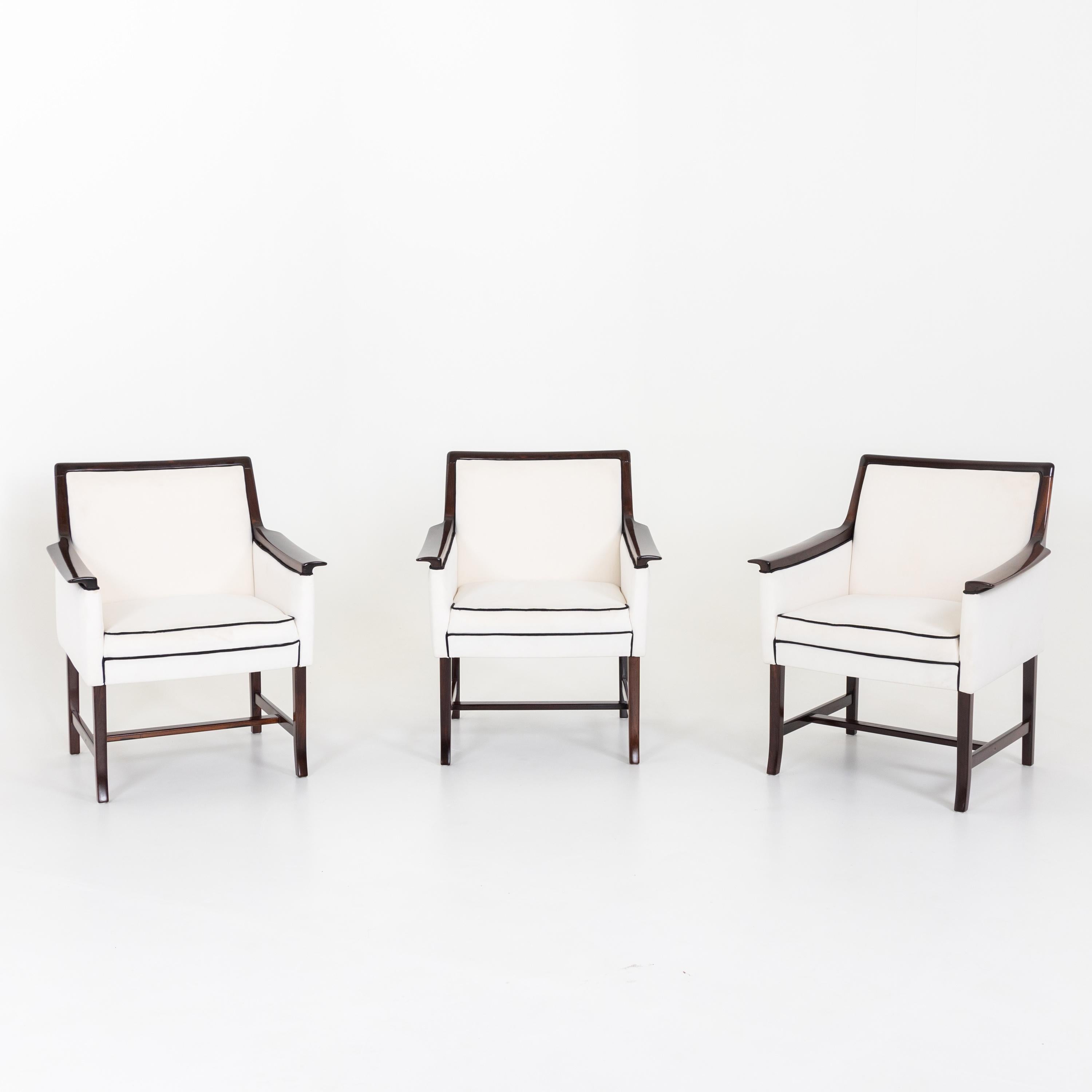 Set of three armchairs on slightly flared legs with H-shaped bracing and upholstered seats with elegant armrests. The chairs have been reupholstered and covered in a white fabric with black piping as well as hand polished.