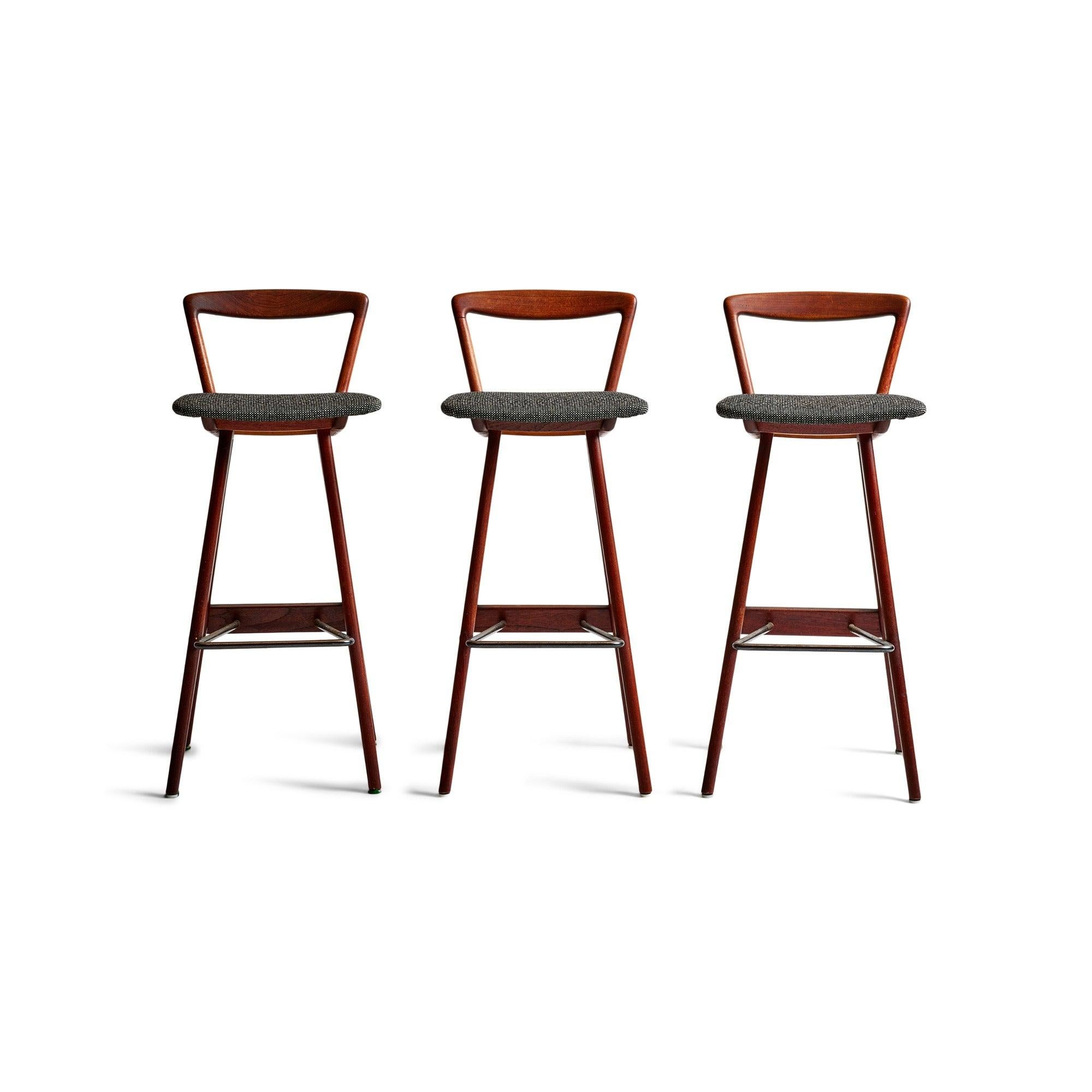 A set of three barstools crafted from teak, featuring cushioned upholstered seats and a tubular metal stretcher serving as a footrest.