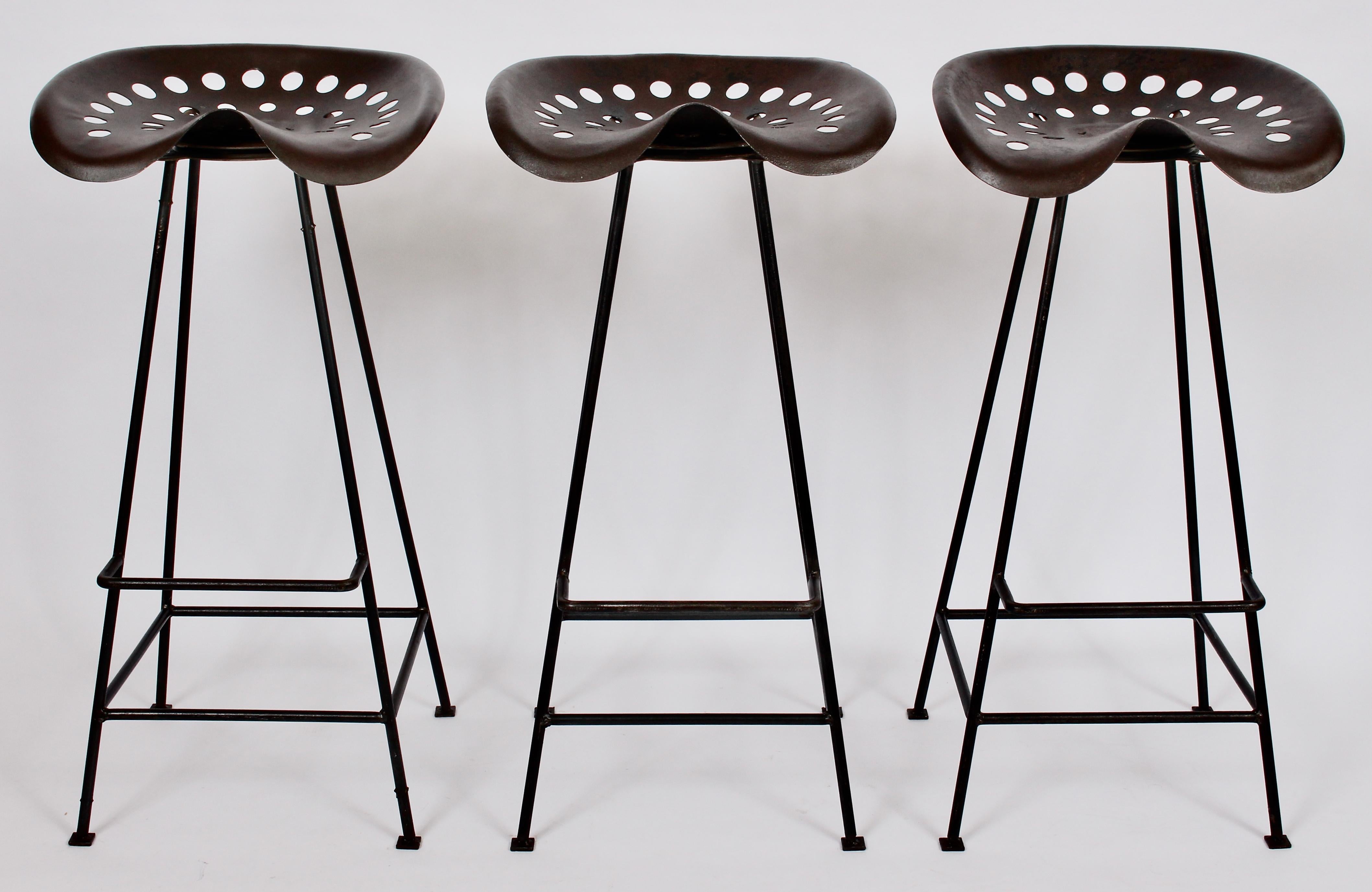 Trio of American Mid Century handcrafted tractor seat bar stools in the style of Benjamin Baldwin. Featuring a sturdy, open Iron framework, footrests with wide perforated patinated and sealed swivel seats (17