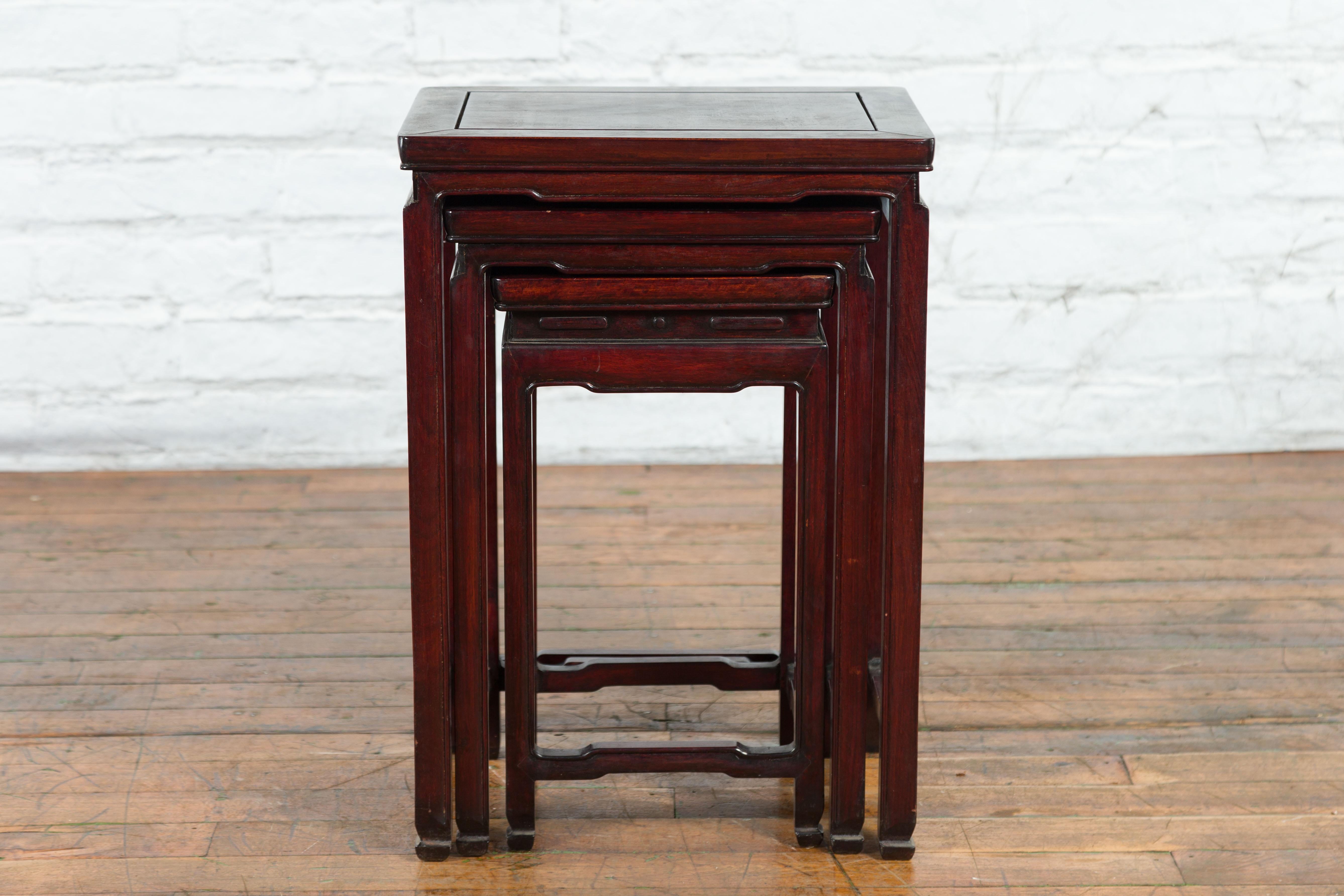 A set of three vintage Chinese rosewood nesting side tables from the mid-20th century with dark reddish brown patina, curving waisted apron, straight legs, horse hoof feet and side stretchers. Created in China during the midcentury period, each of