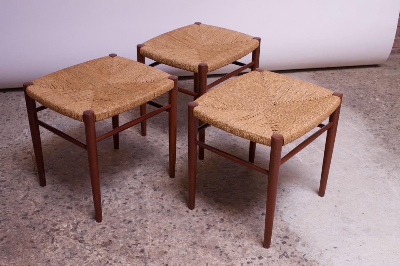 Set of three circa 1950s Danish Modern low / vanity / foot stools with woven paper cord seats and solid teak frames. The teak has been refinished; the rush is in good, vintage condition with minor wear, as shown. 
Measures: H 18