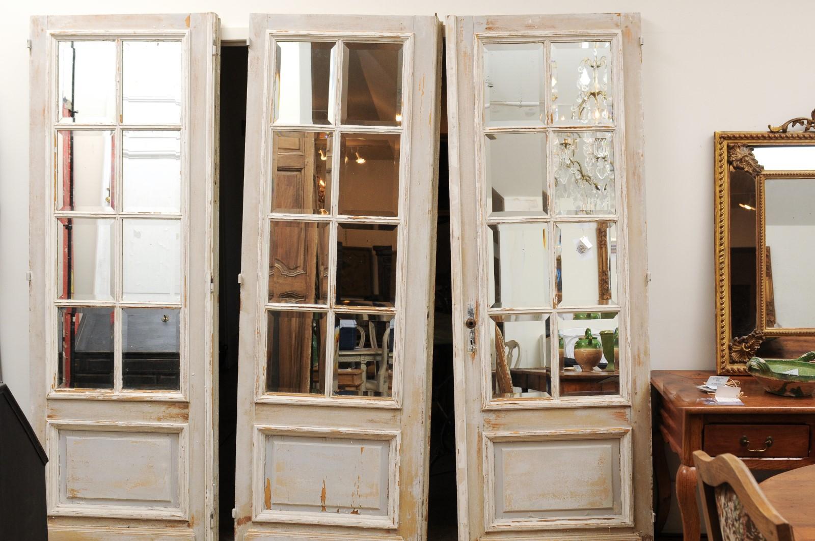 A set of three French painted wood mirrored doors from the 20th century, with nicely distressed patina. Created in France during the 20th century, each of this set of three doors features a linear frame perfectly accented with mirrored panels in