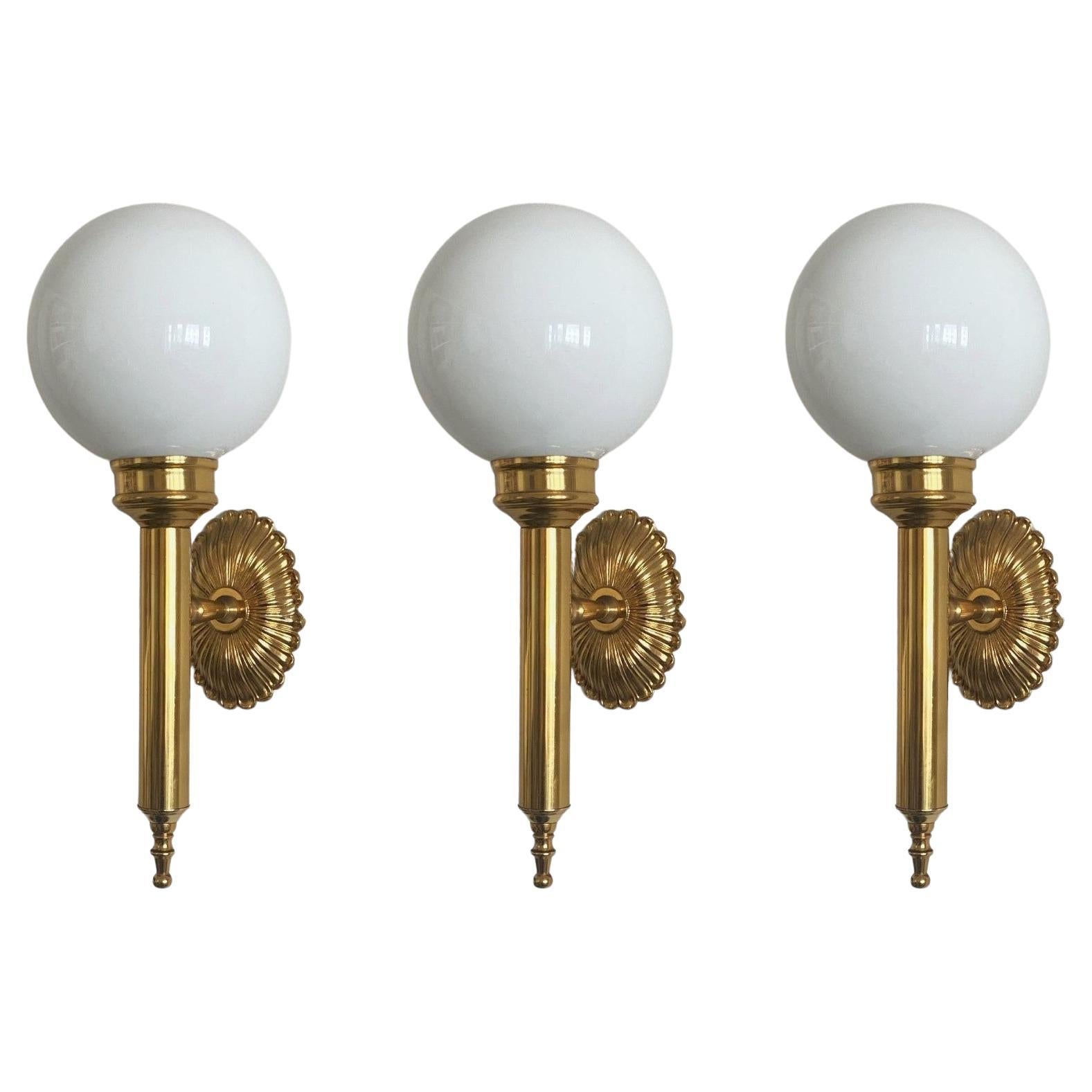 A lovely set of three Art Deco torchiere wall lights, France, 1930s. Elegant design in gilt brass with hand-blown opaline glass ball globes. 
All three pieces in fine vintage condition, glass globes without chips or cracks, aged patina to brass,