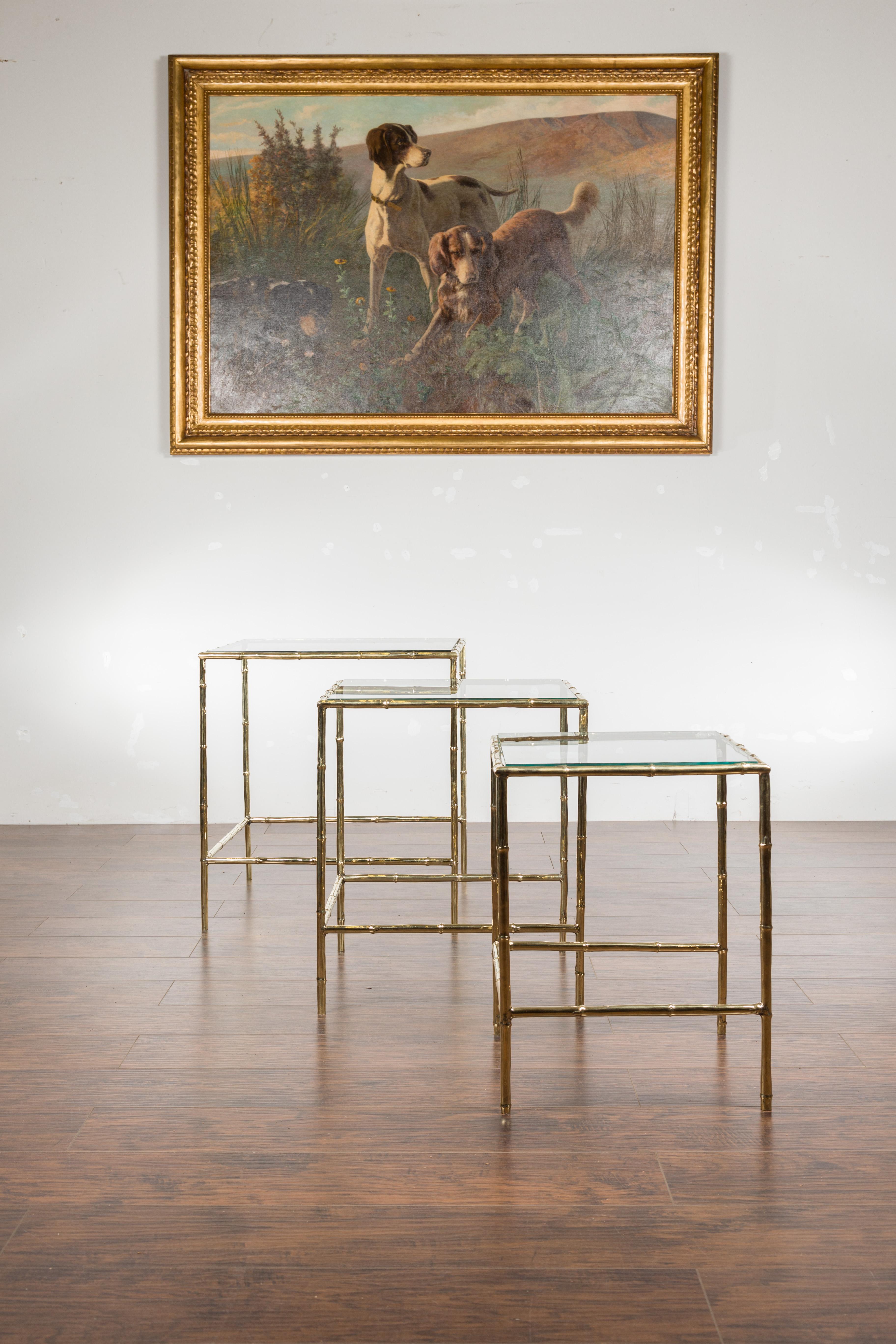 A set of three French vintage brass faux-bamboo nesting tables from the mid-20th century, with glass tops and stretchers. Created in France during the mid-century period, each of this set of three 