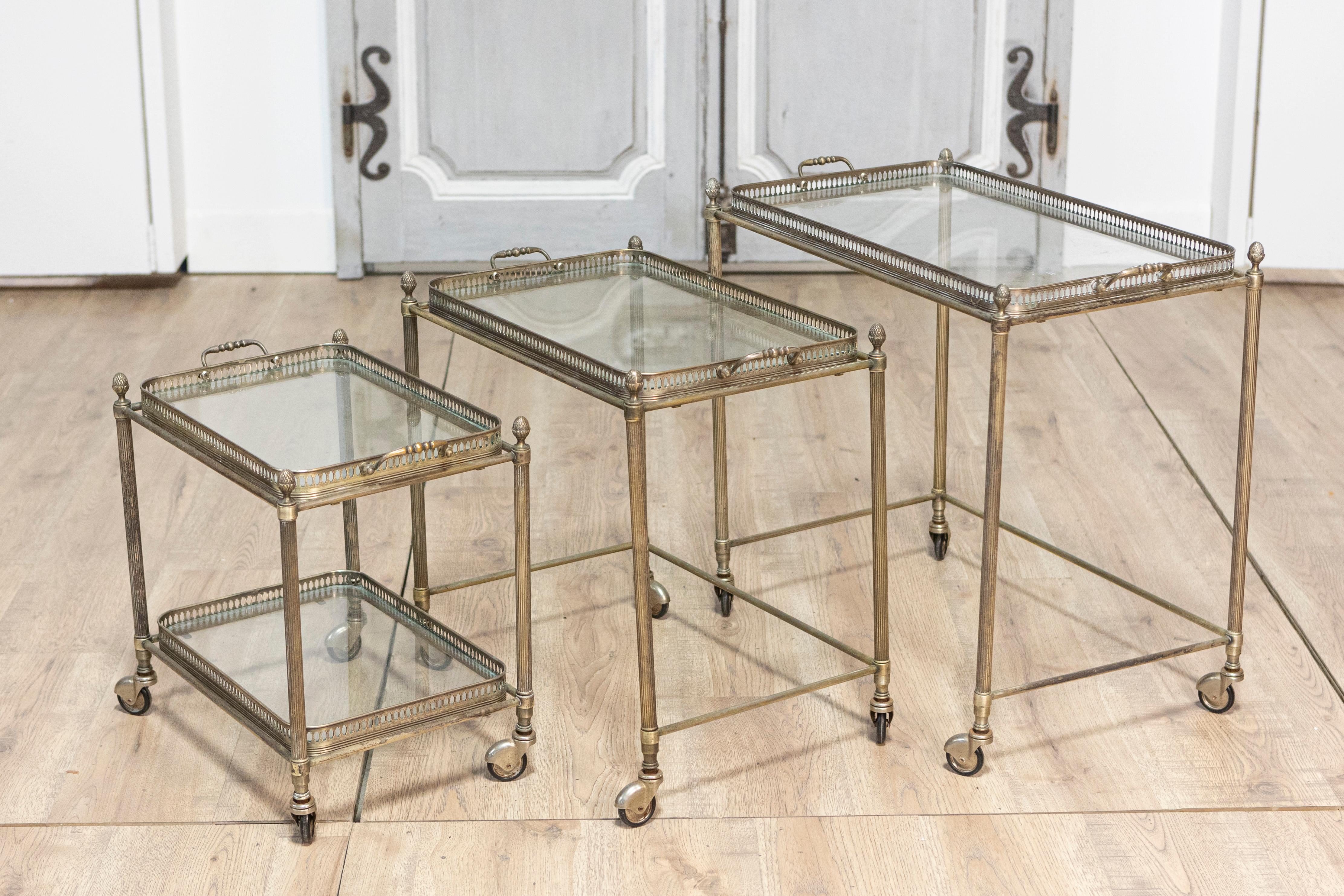 Un ensemble de trois chariots de bar gigogne de style néoclassique français du 20e siècle avec des galeries percées, des étagères en verre, des épis de faîtage en forme de gland et des pieds cannelés sur roulettes. Cet élégant ensemble de trois
