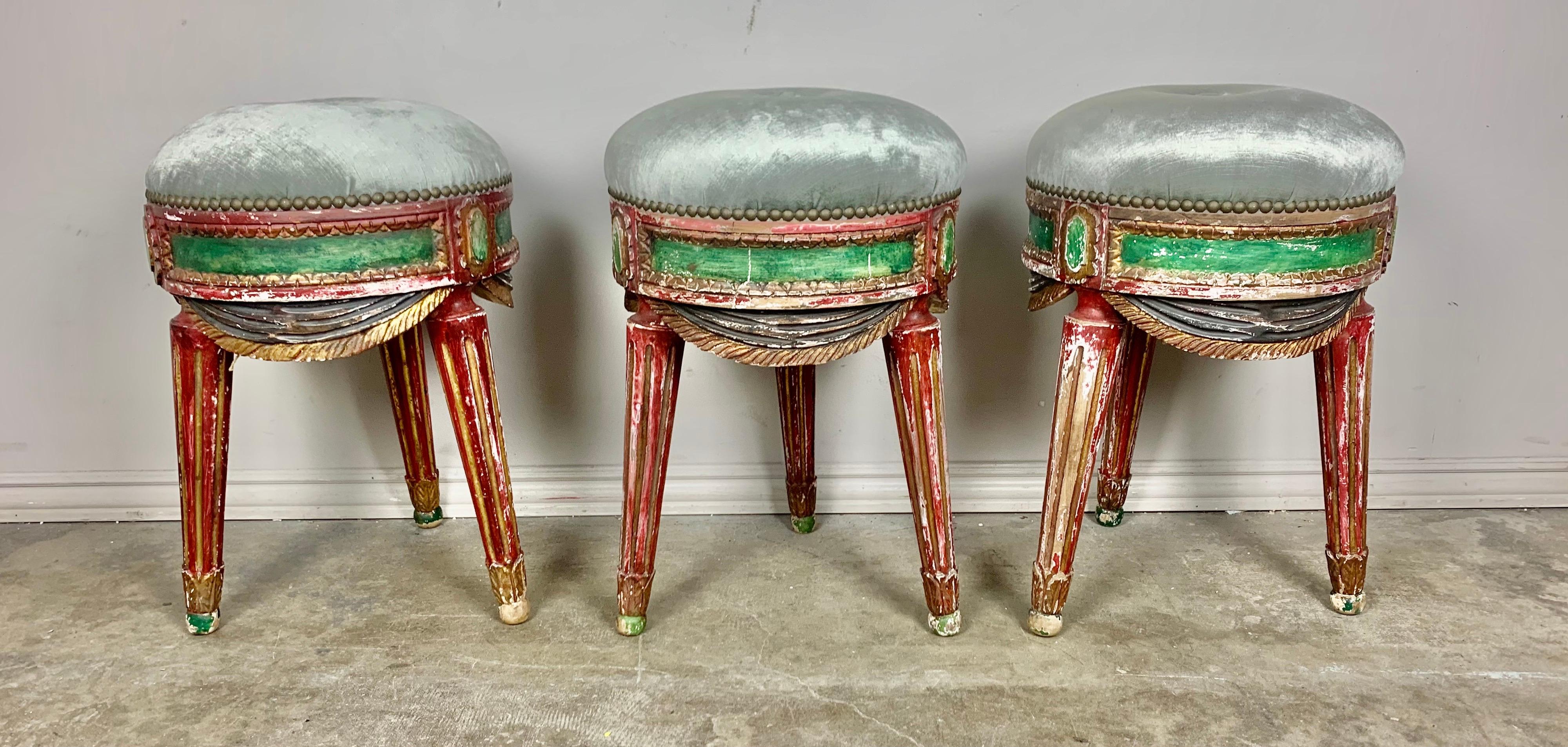 Set of three carved wood painted tripod stools standing on three fluted legs with celadon velvet upholstery. The benches are painted in vibrant colors of red, green & blue. The upholstery is finished with nailhead trim detail. Great carved details