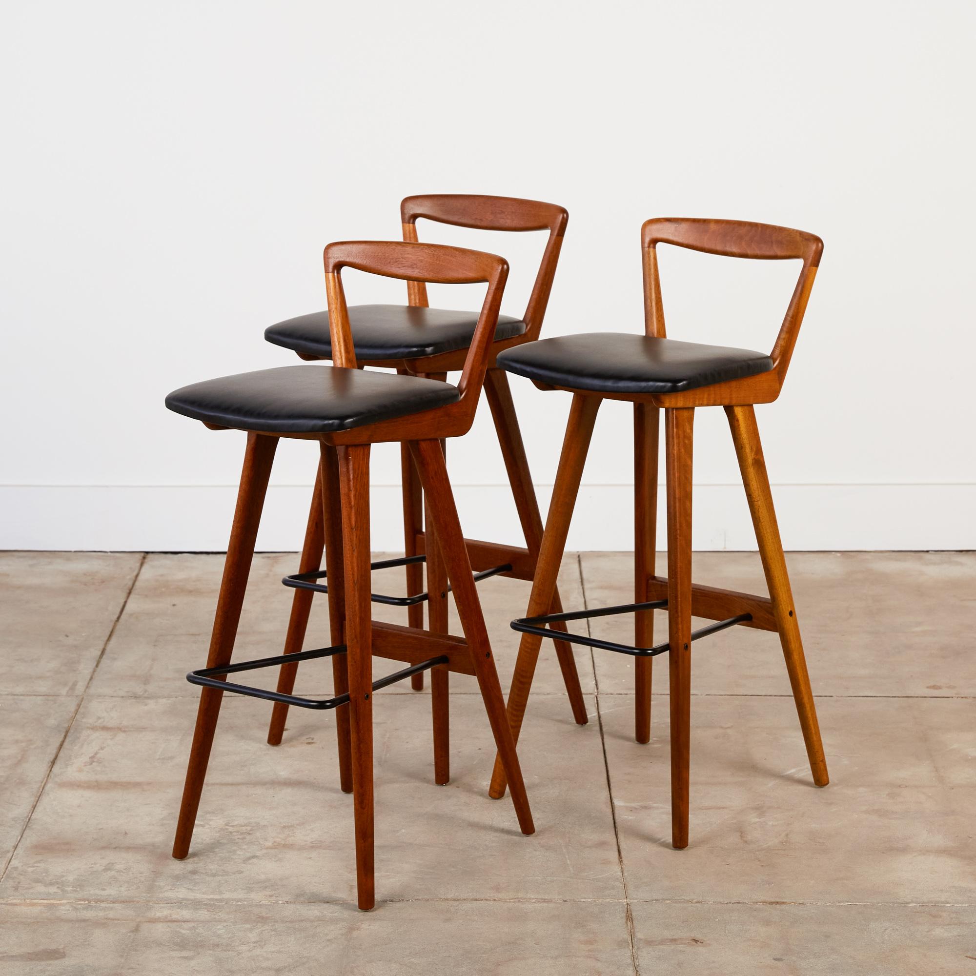 A set of three 1960s bar stools by Henry Rosengren Hansen for Brande Møbelindustri. The stools have new black leather upholstered seats and sit on frames of solid teak. Each stool has flat, tapered legs and an open backrest. The horizontal