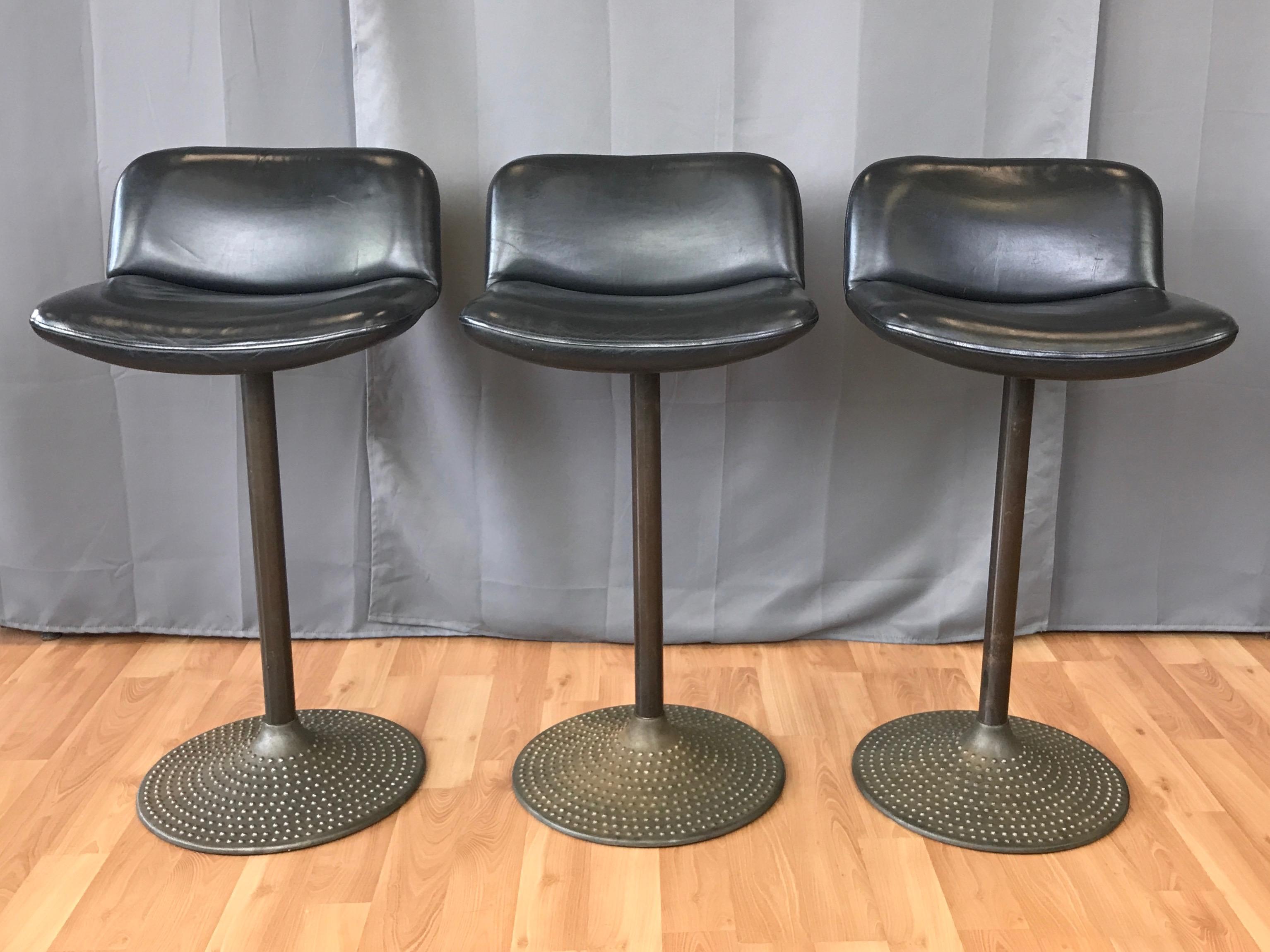 A set of three Caribe swivel barstools with leather seats by Ilmari Tapiovaara for ICF.

Hobnail textured cast steel base and tubular stem have an understated patinated bronze finish. Broad swivel seat with low back features original black leather