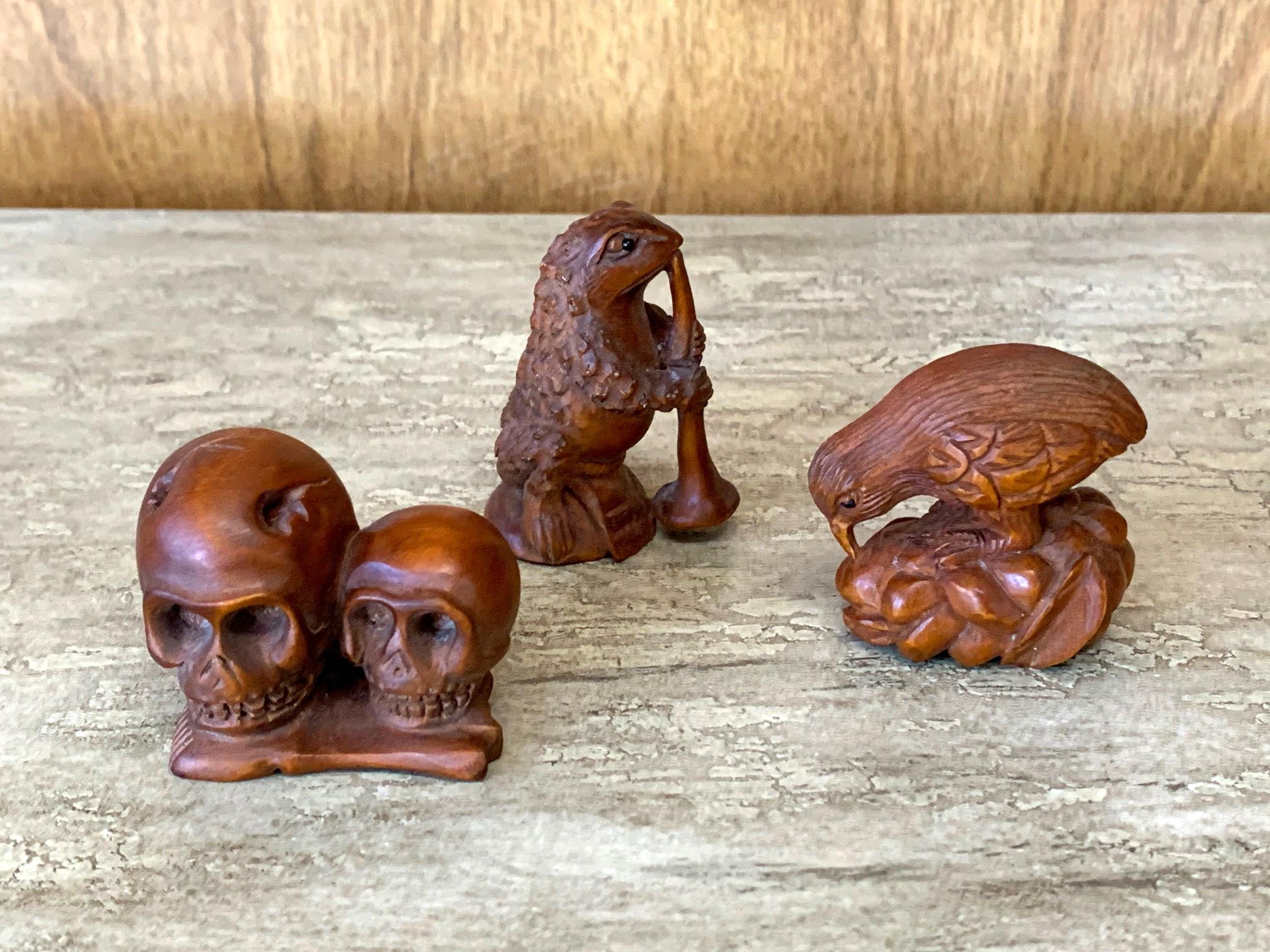 Set of three Japanese netsukes carved out of wood with a reddish stained finish, appear to be circa mid-20th century. One depicts a frog holding a trumpet; one a rooster eating grains, one two skulls with base made of bone. None signed. Sold as a