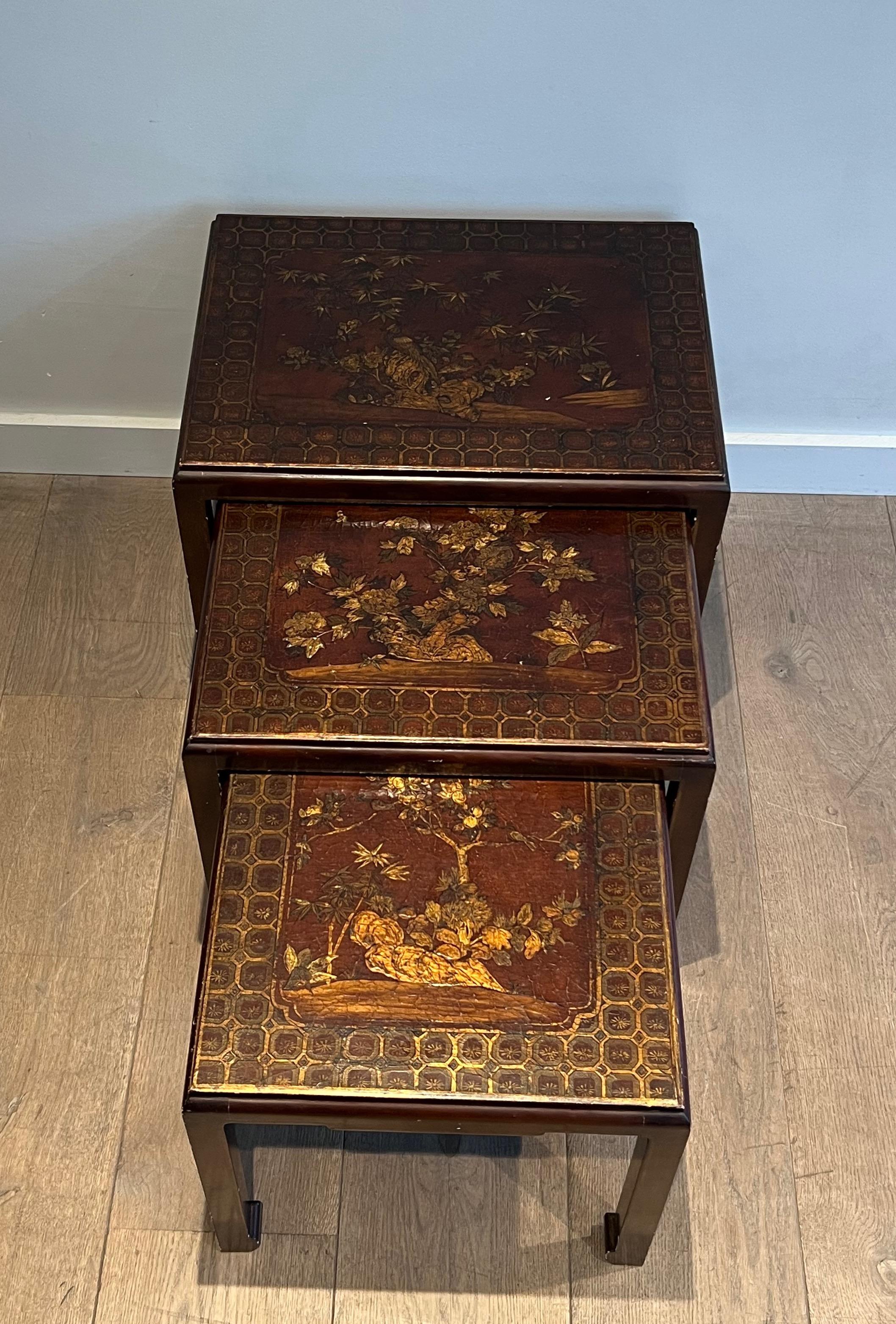 This very nice set of three nesting tables is made of a lacquered wood in the bordeaux tones with gilt decors representing Chinese scenes. This is a French work. Circa 1940.