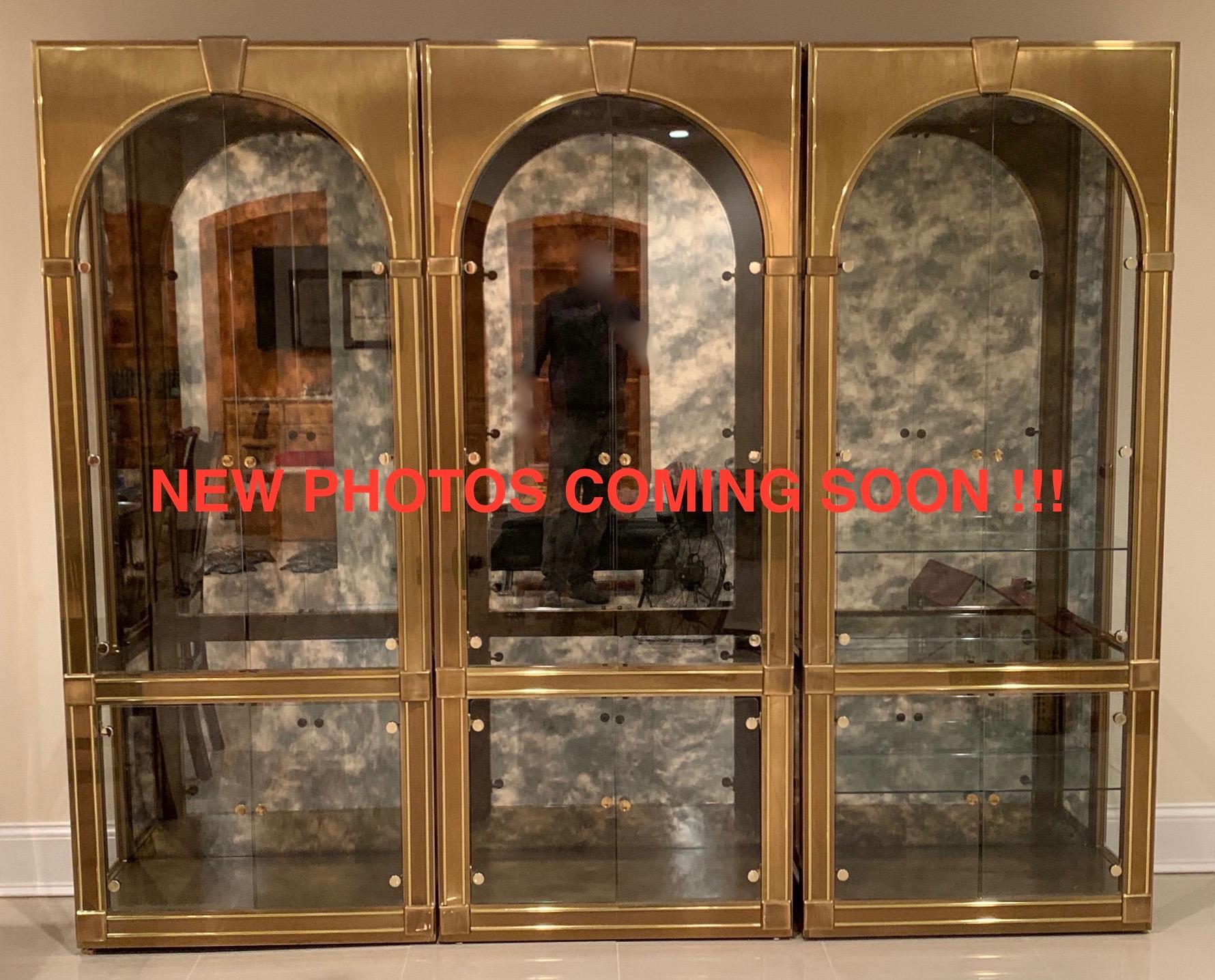 Set of Three Glamours 1970s Mastercraft Palladian style brass clad display cabinets or vitrines with arched doors and keystone pediment. The lighted interior has five glass shelves, four that are adjustable, etched plate grooves and a dramatic