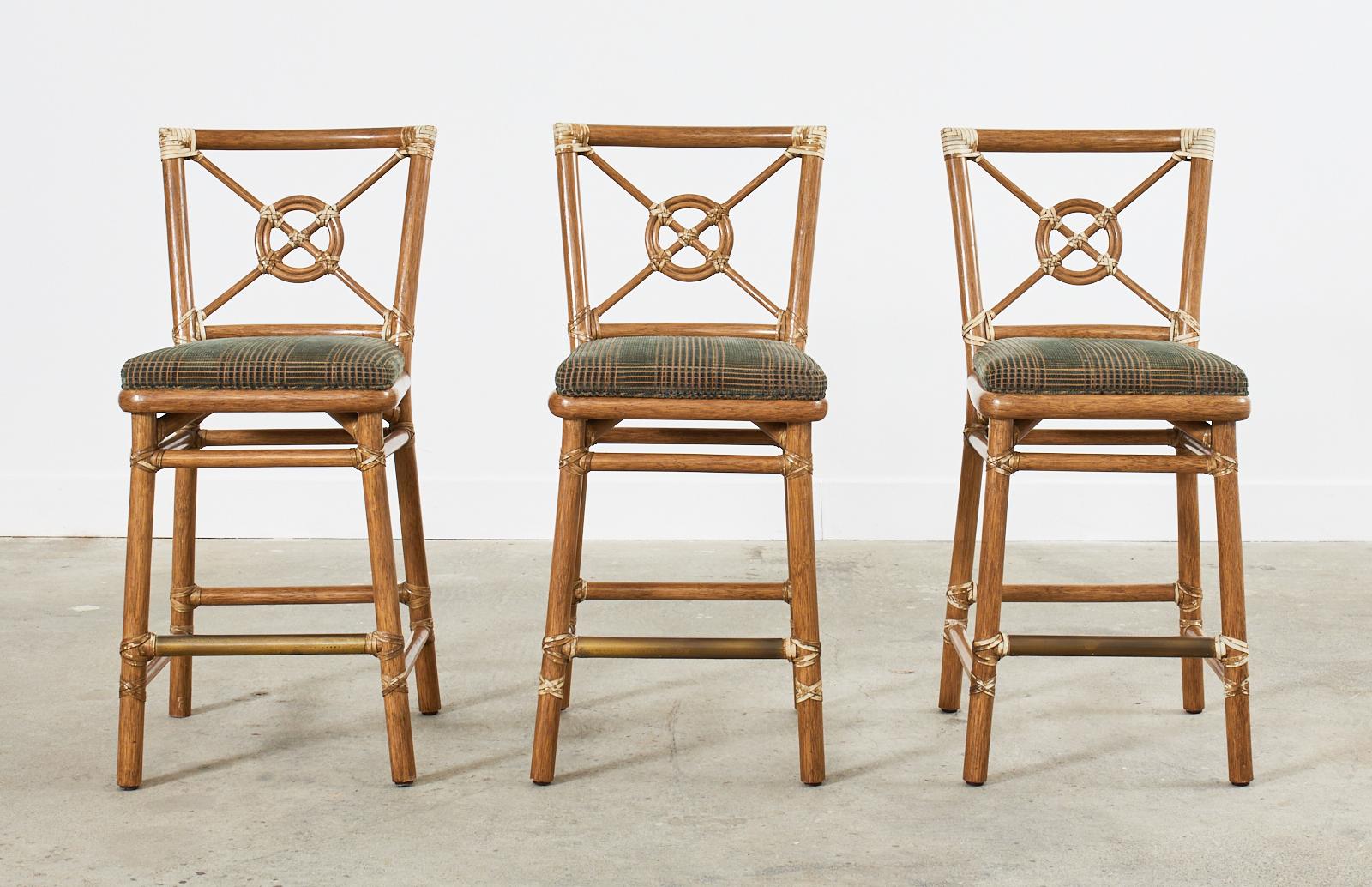 Iconic set of three McGuire rattan counter height barstools featuring the target design motif designed by Elinor McGuire. The thick rattan frames are lashed together with leather rawhide laces. The stools have a footrest in the front with an