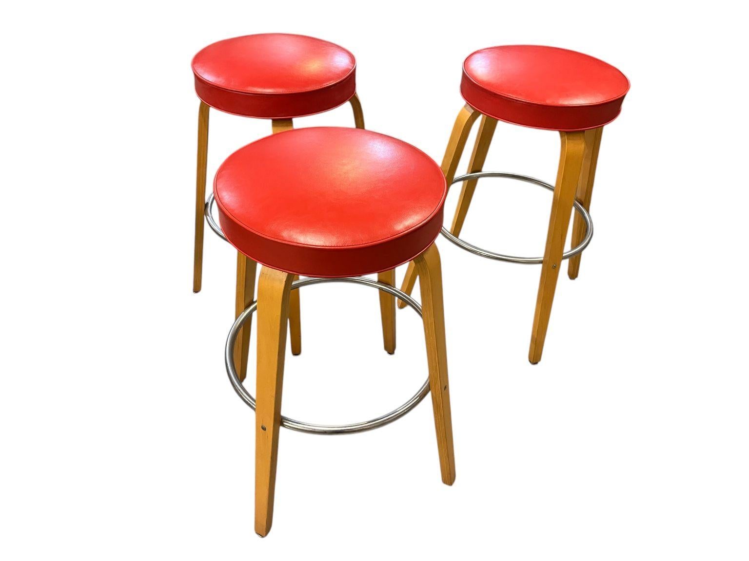 Set of three Thonet bar stools in a light blonde maple with red Naugahyde vinyl with a round chrome foot rest. In beautiful original condition with an original tag on one of the stools. Tag states Thonet Industries, One Park Ave NY, NY. Dimensions