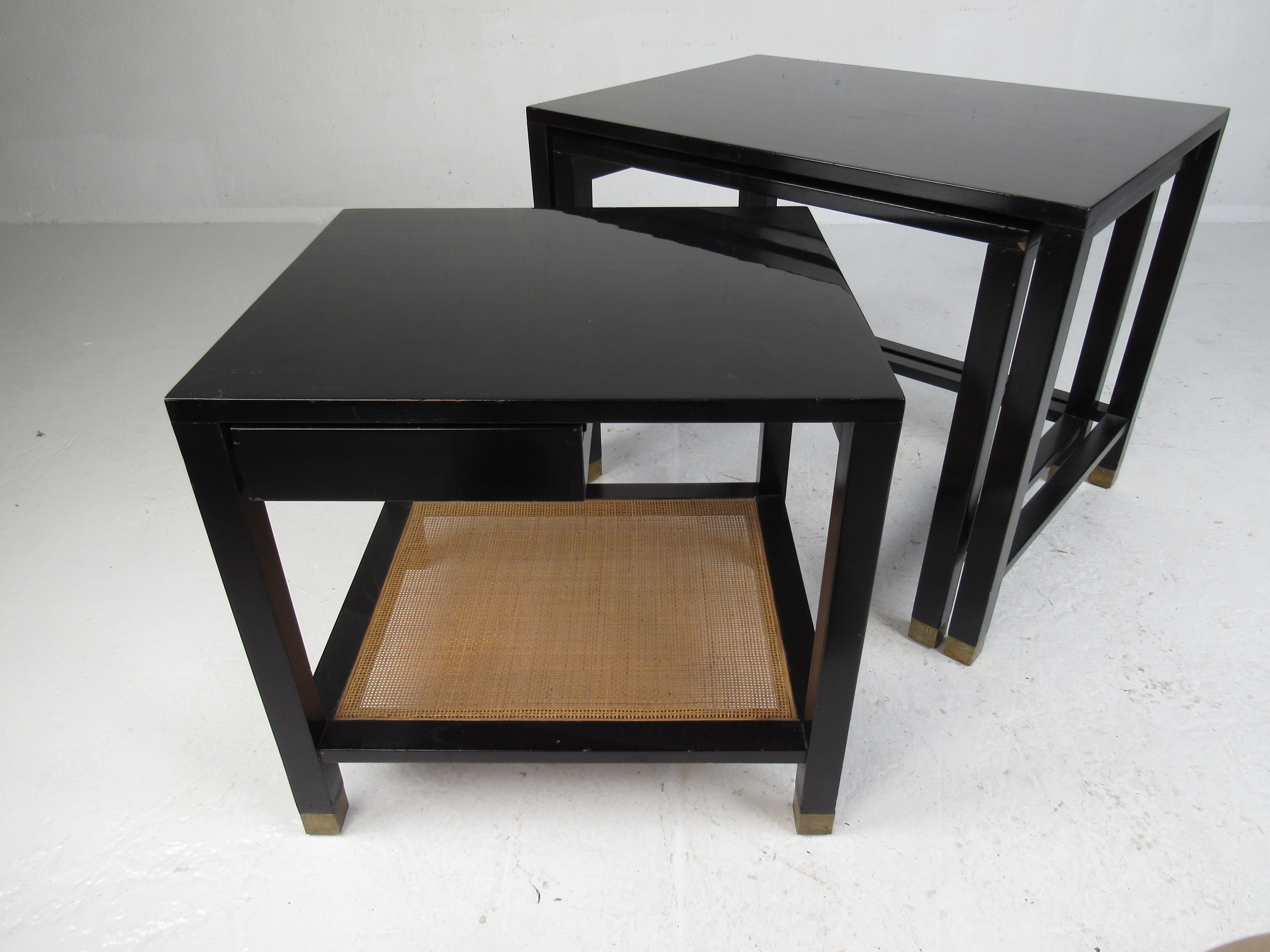 A stunning set of three vintage modern nesting tables with a black lacquered finish and brass capped feet. The smallest table boasts a conveniently placed drawer and a unique lower cane shelf ensuring plenty of room for storage. These beautiful