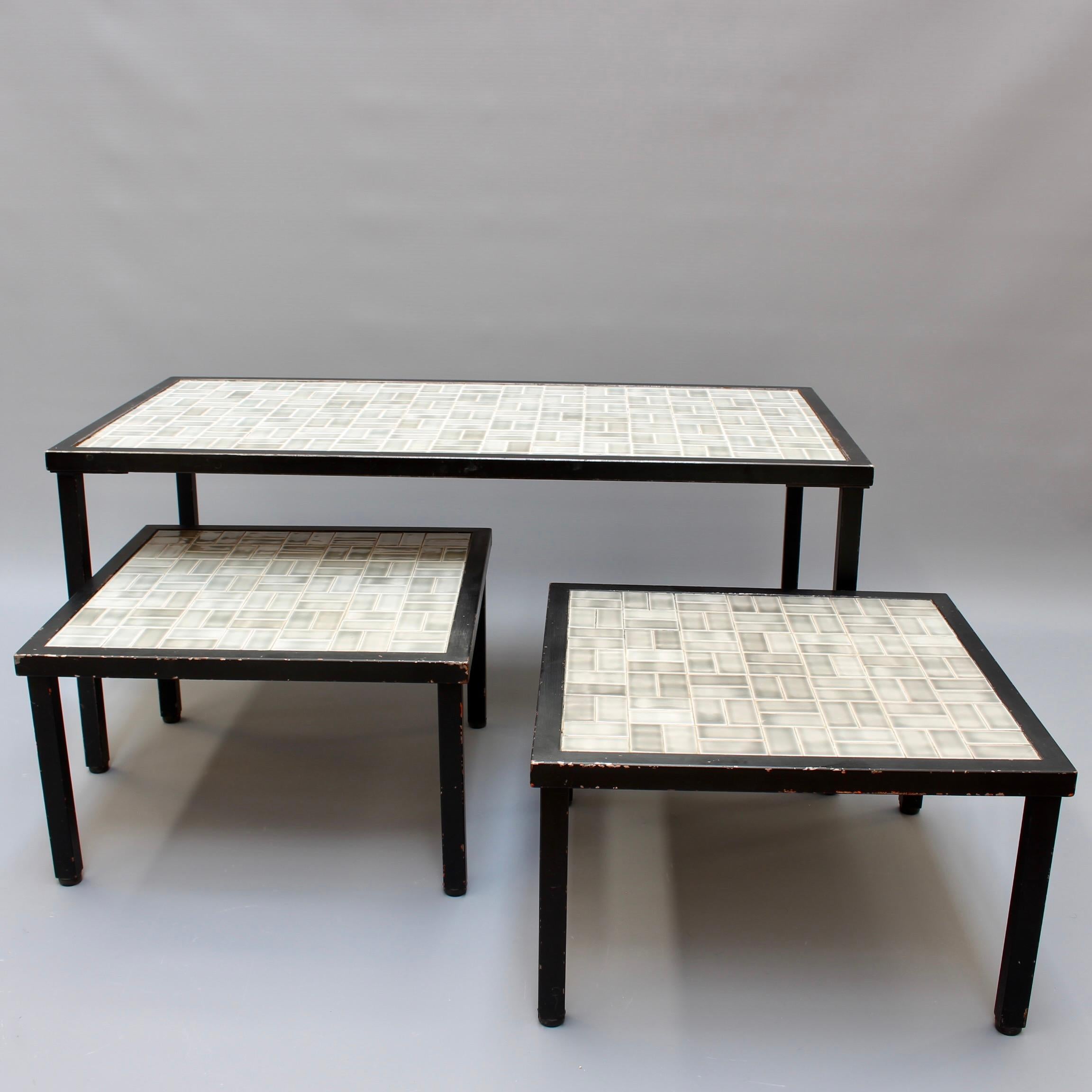 Set of three vintage French tiled coffee and end tables (circa 1960s). Subdued hues shade the ceramic tiles which sit upon simple frames and black metal legs in modern symmetry. The tiles are grey, cream and a faded black seemingly painted with