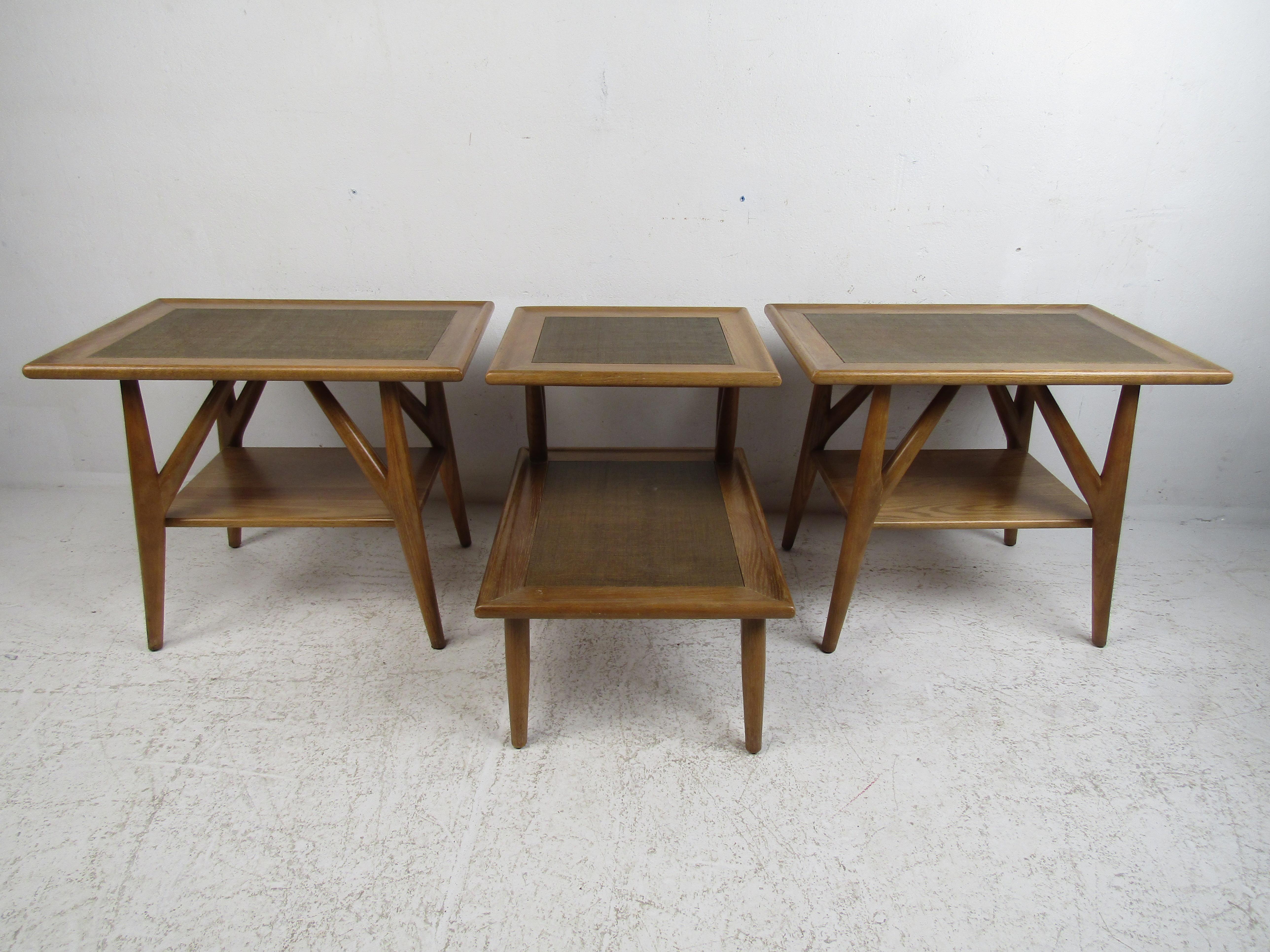 This unusual set of three vintage modern end tables boast solid oak frames with grass cloth tops. A gently used set with unique stretchers that angle upward, splayed legs, and slightly raised edges along the top. A rare step-side table included in
