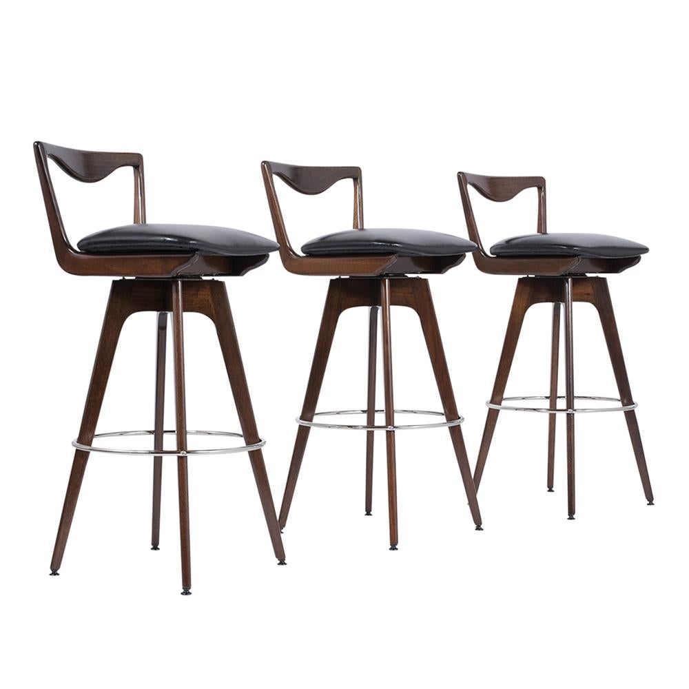 These three midcentury leather bar stools are made out of walnut wood, feature a curved back design, and rest on stretched legs frame newly stained and lacquered finish. The seats have all been upholstery in new black leather with new comfortable