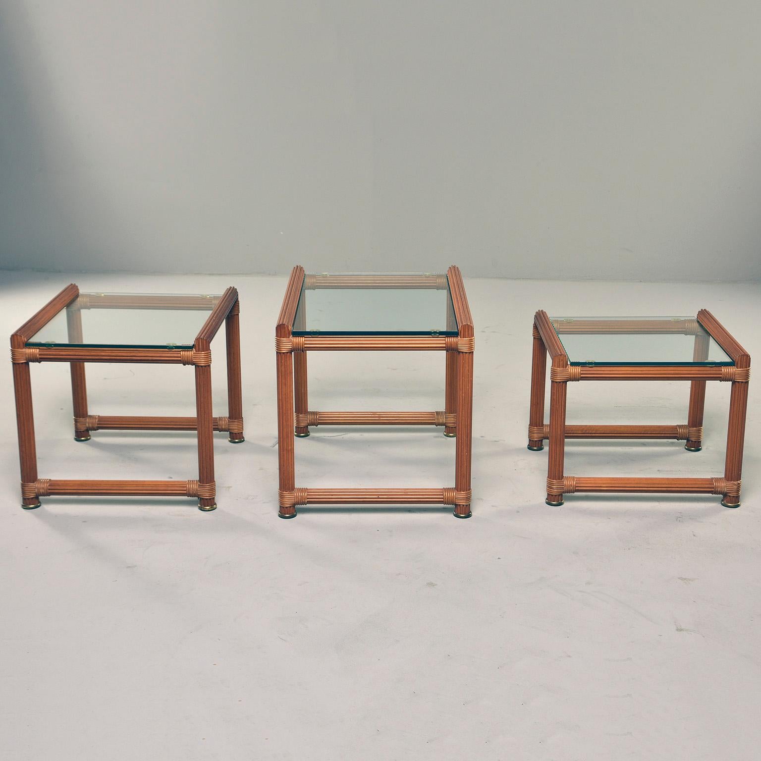 Trio of nesting tables feature reeded wood frames with brass rimmed feet and glass tops, circa 1970s. Found in Italy. Unknown maker. Sold and priced as a set of three. Measurements of three tables are as follows:

Large 17.5” H x 23” W x 17”