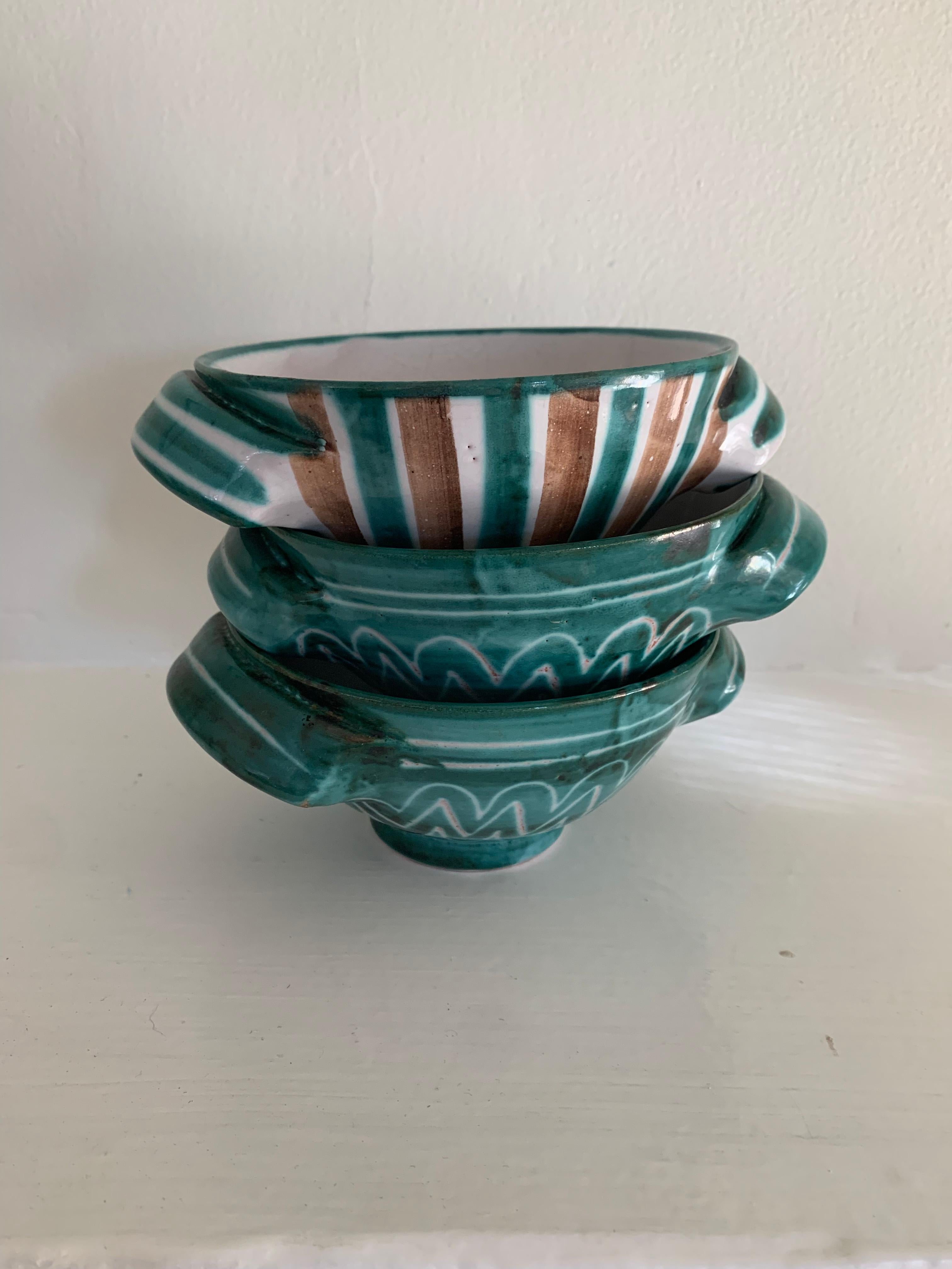 Descrizione: Set di tre ciotole con manico in ceramica di Robert Picault, realizzate a mano e decorate con i suoi smalti turchesi, marroni e bianchi. Due presentano motivi a zig zag e uno a strisce verticali alternate, che rivelano la calda