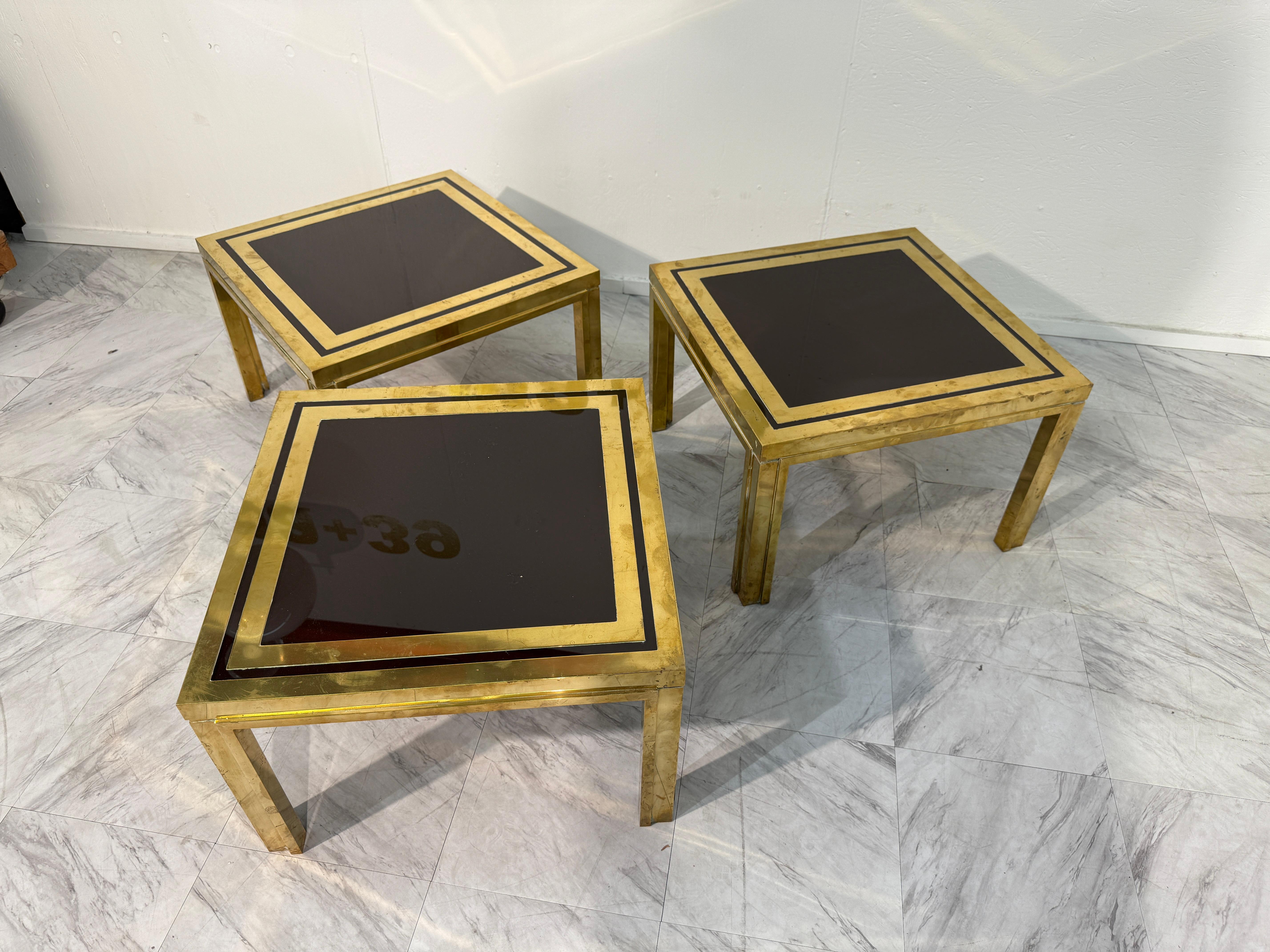 Un iconico set di tre tavolini quadrati italiani di Romeo Rega, disegnati negli anni '70. Ogni tavolo presenta un'audace struttura in ottone dalle linee geometriche pulite, abbinata con eleganza a un piano in vetro fumé accentuato da un doppio bordo