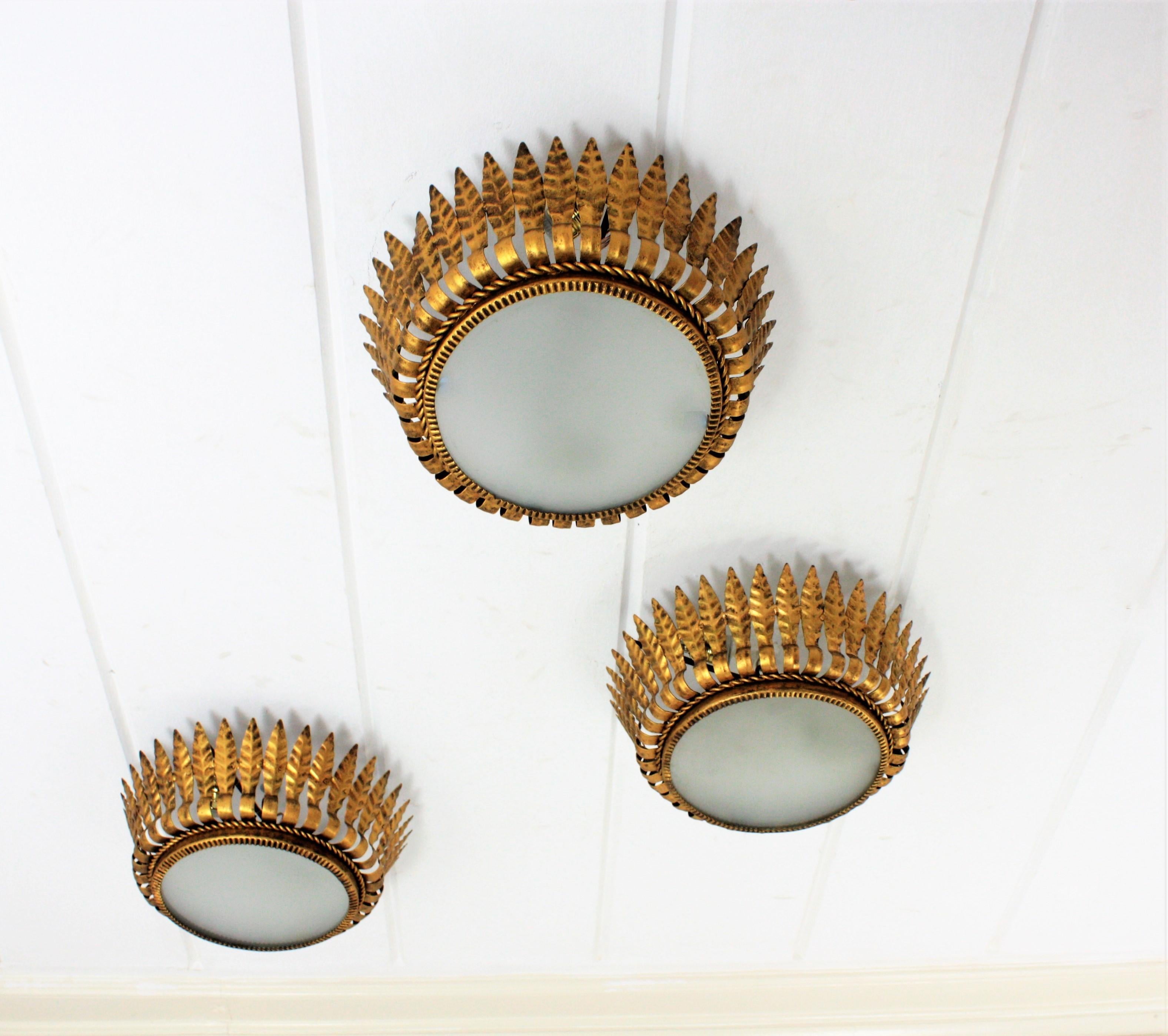 Set of three Spanish Mid-Century Modern gilt iron crown sunburst light fixtures or chandeliers with leaf motifs surrounding a central frosted glass difusser, Spain, 1950s.
All of them have been handcrafted in hand-hammered iron with gold leaf