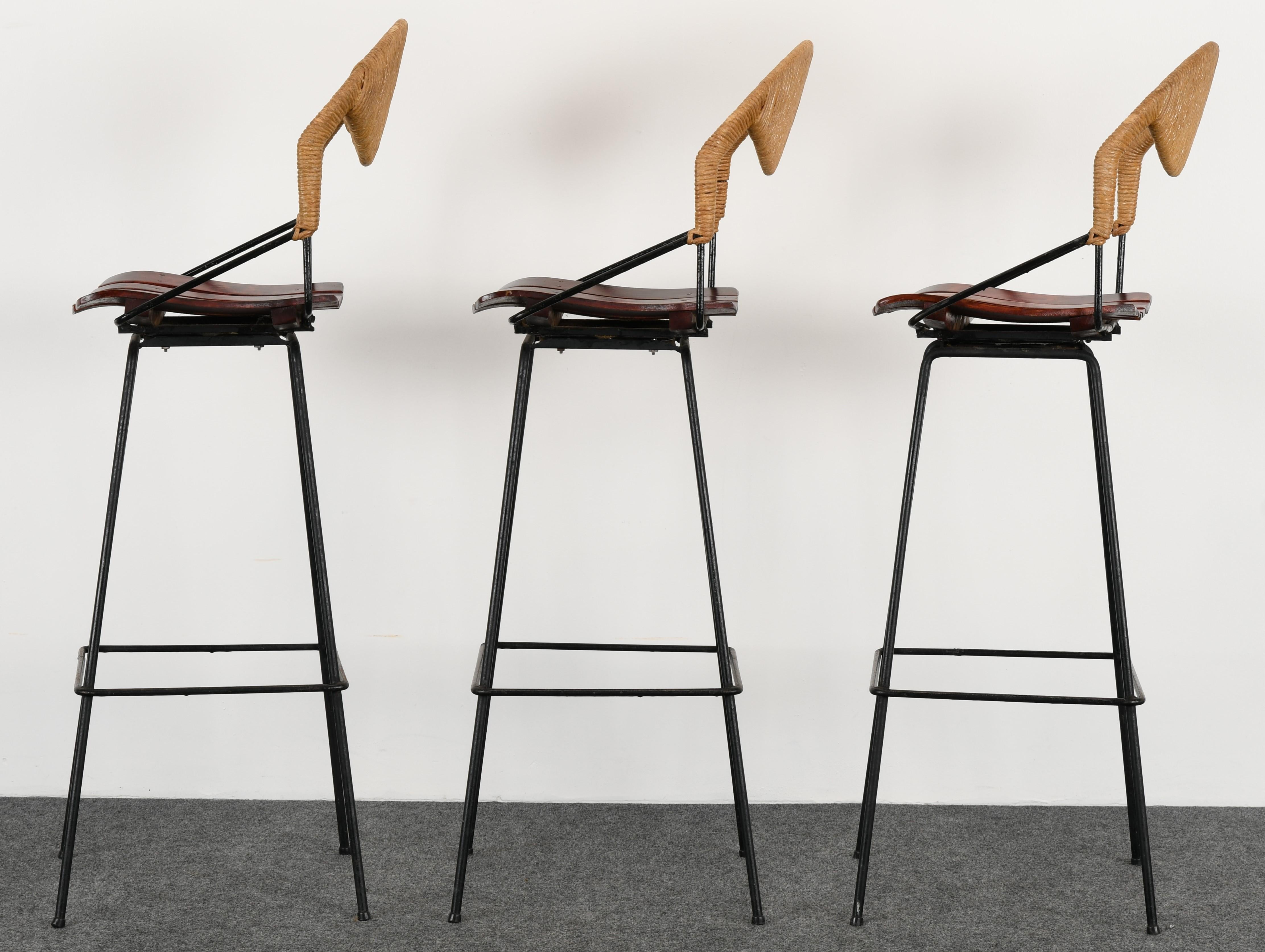 A set of three swivel bar stools by Arthur Umanoff, 1960s. The stools have wood slat and rushed back seats with black metal iron base. Stools are in good condition with minor scratches to rush but not distracting.

Dimensions: 45.5