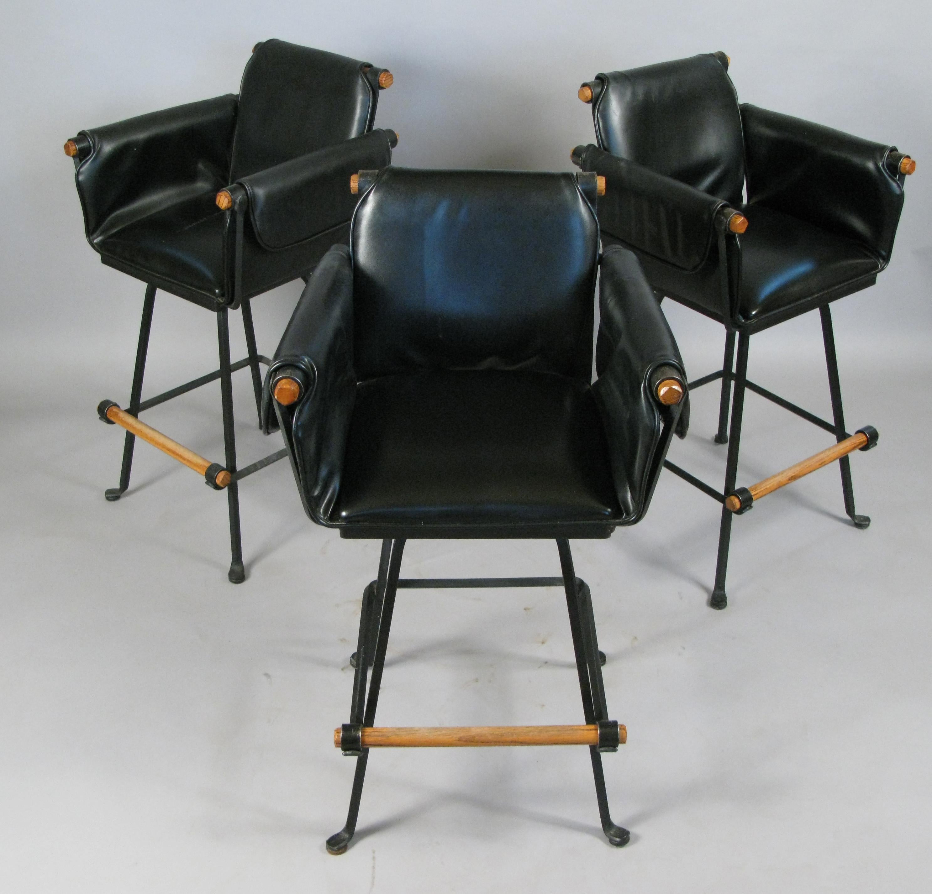 A beautiful set of three vintage 1970s swivel barstools with wrought iron frames and oak arms and footrests. With their original black vinyl upholstered seats and arms. Labeled Terra Furniture.