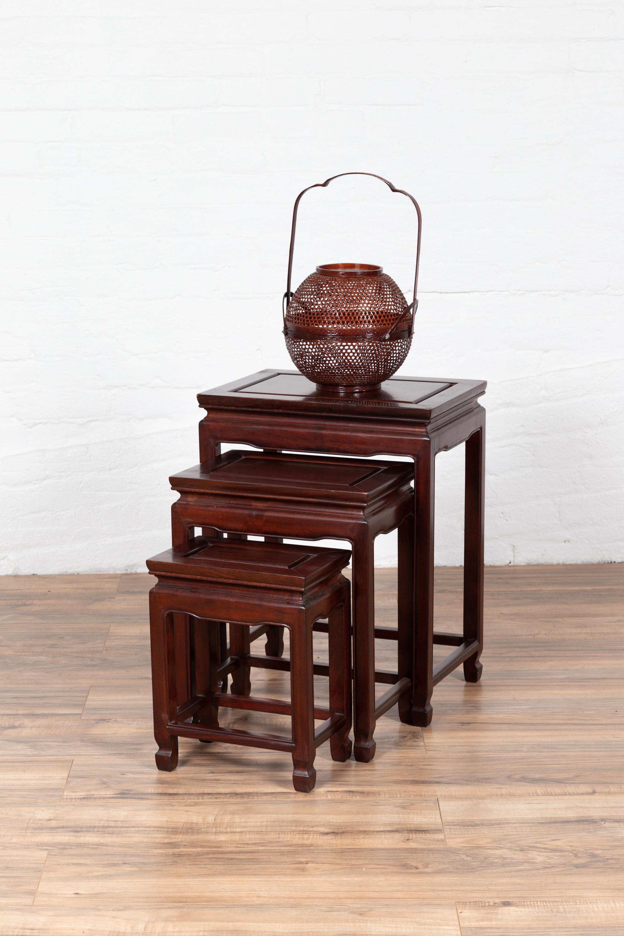 A set of three vintage Chinese rosewood nesting side tables from the mid-20th century with dark patina, curving waisted apron and straight legs. Born in China during the mid-century period, this exquisite set of rosewood nesting tables each features