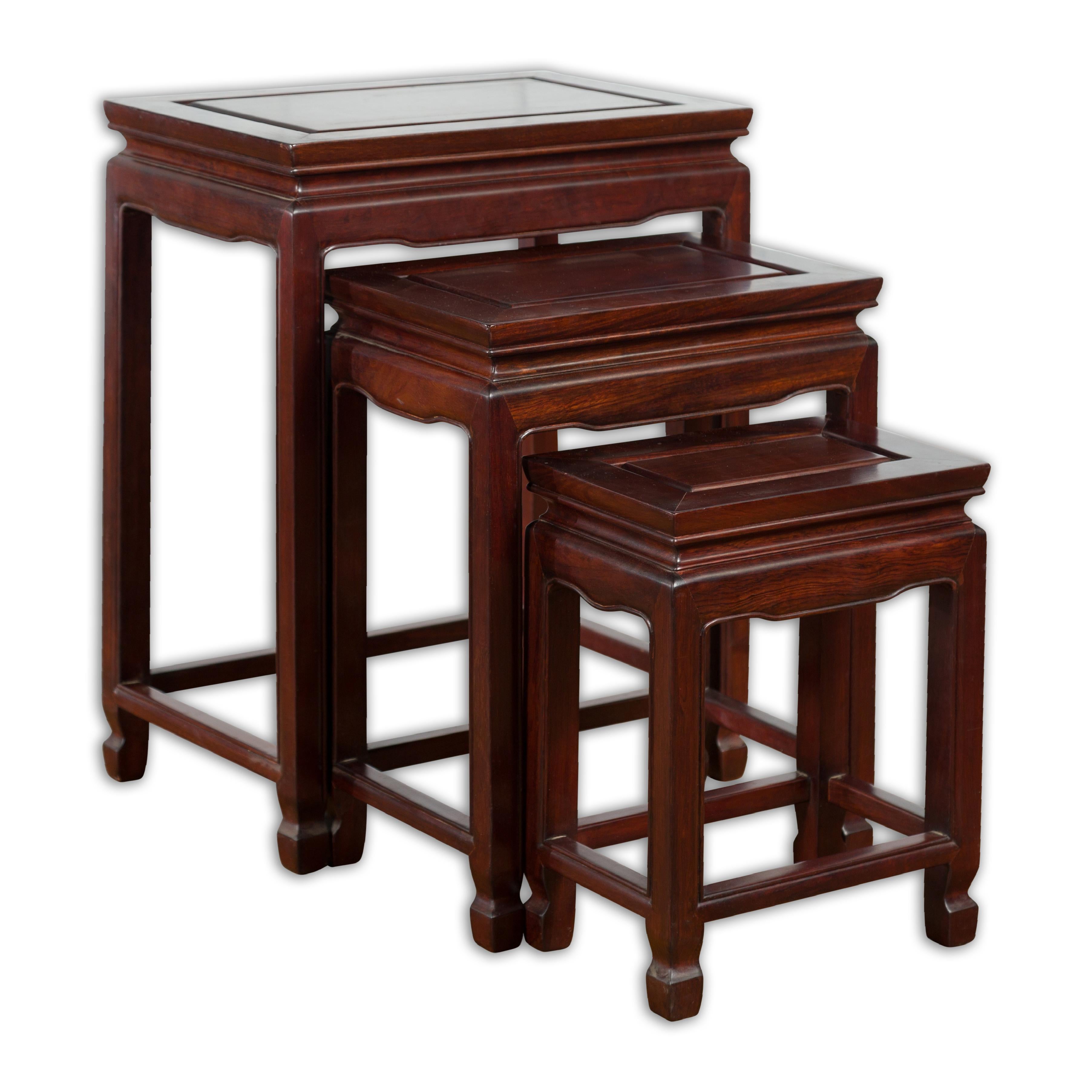 A set of three vintage Chinese rosewood nesting side tables from the mid-20th century with dark reddish brown patina, curving waisted apron, straight legs, horse hoof feet and side stretchers. Created in China during the midcentury period, each of