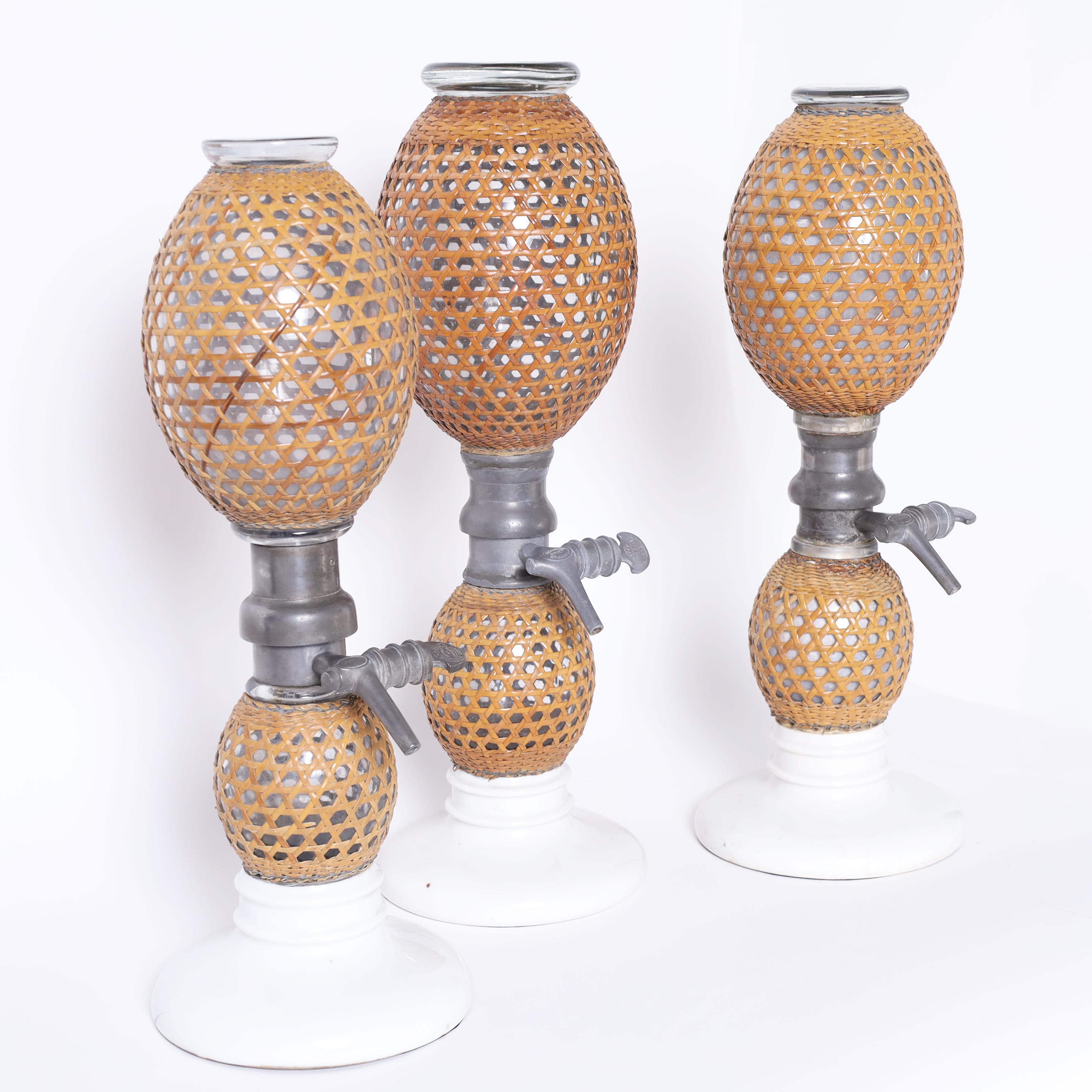 Transporting set of three French seltzer bottles with iconic form crafted with hand blown glass bulbs wrapped in reed having pewter hardware and porcelain bases. 

From left to right:

H: 17.5 DM: 6.5
H: 18.5 DM: 6.5
H: 18 DM: 6.5
