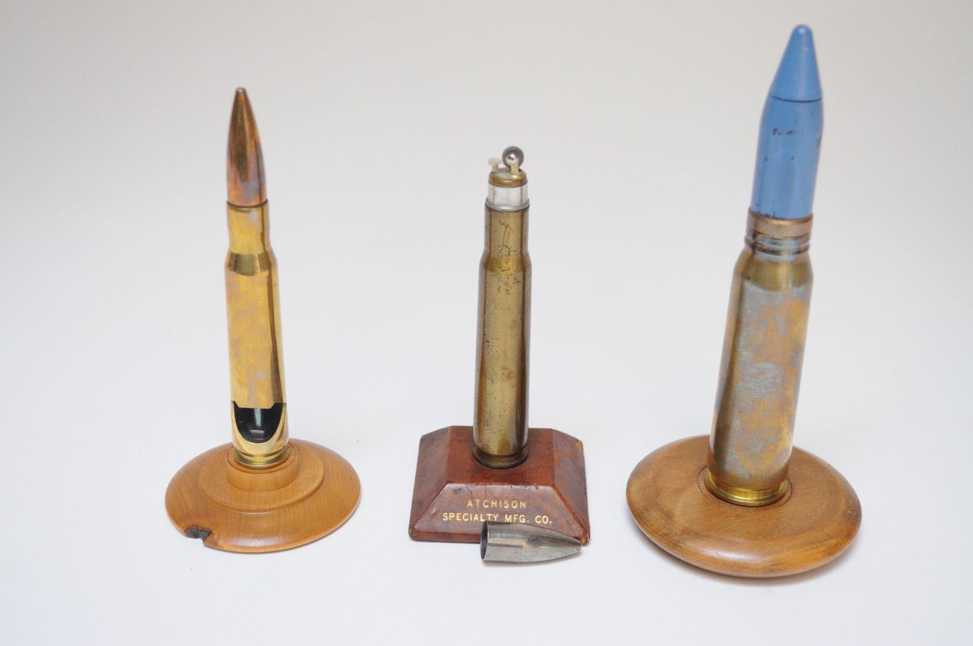 Collection of three trench art figurines composed of gutted shell casings mounted to bases. Two are decorative paperweights supported by round, wooden, platform bases, while the third, an actual flint lighter concealed by the projectile, is