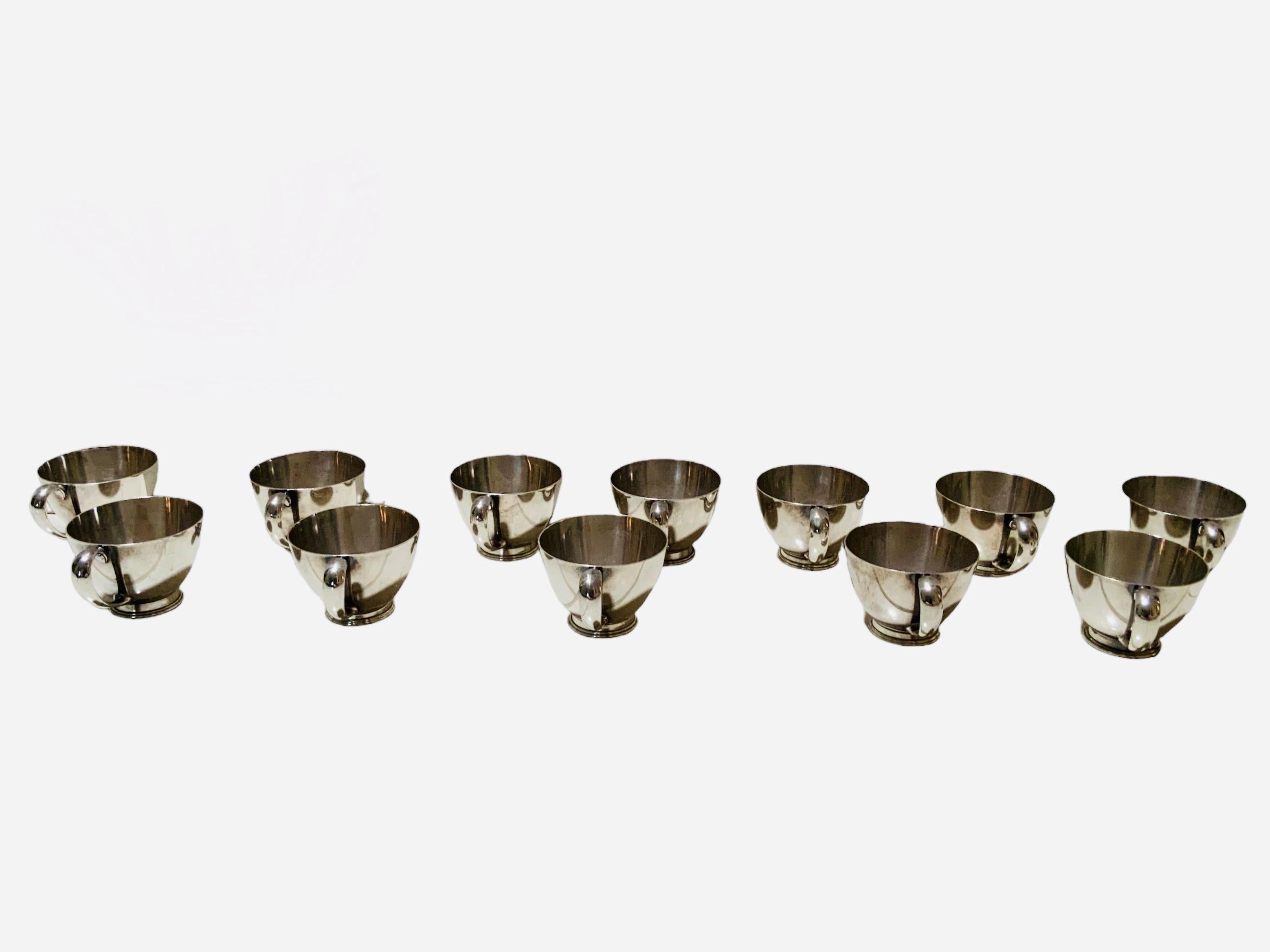 This is a set of twelve Cartier sterling cups. The cups have wide mouths and flared handles.They are adorned with a ribbed round pedestal. Below them, it is found the Cartier hallmark with GWO shields mark and Sterling 224 inscribed.