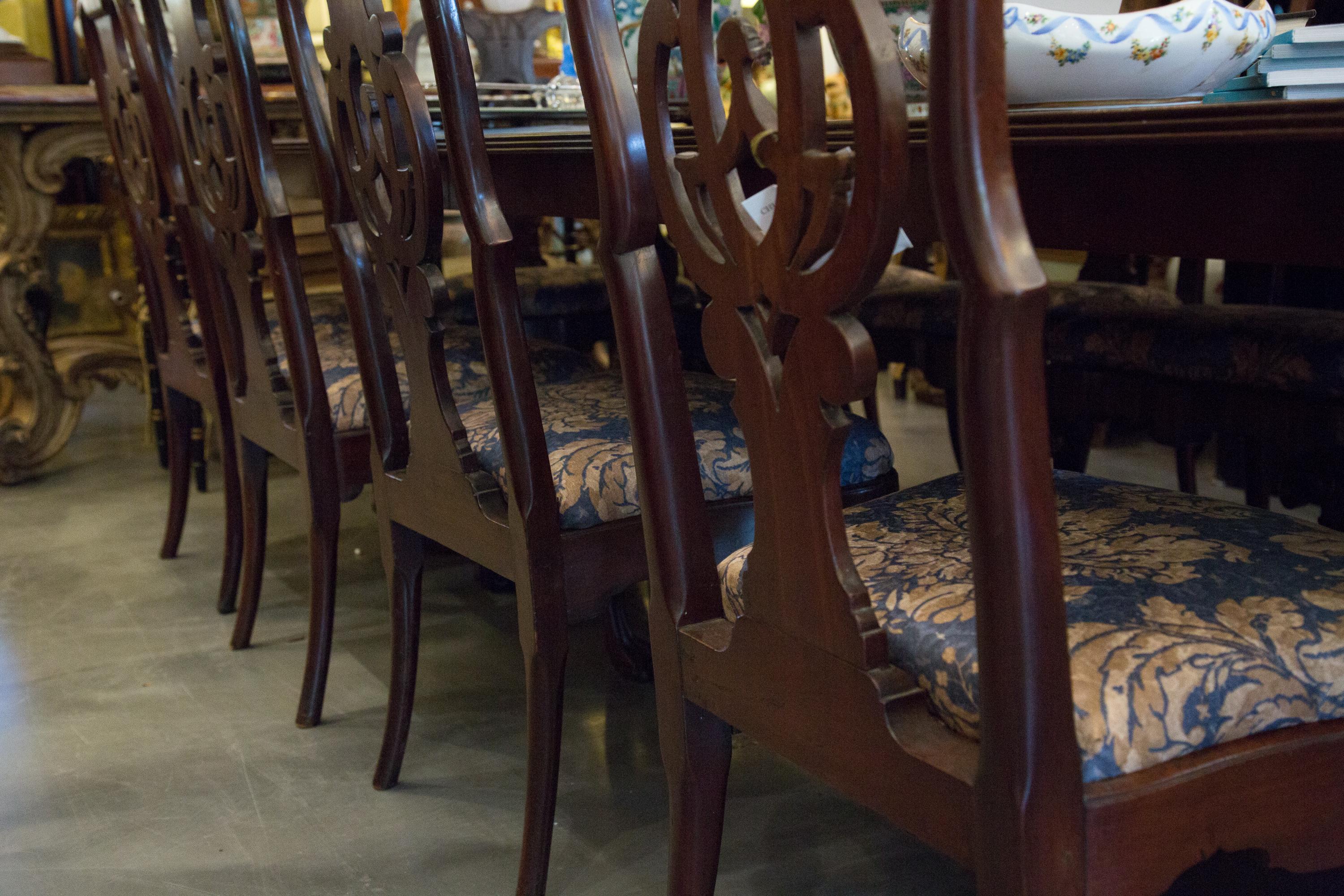 This is a lovely set of 12 George II style mahogany dining chairs, consisting of two armchairs and 10 side chairs. Each chair has an intricately carved pierced back slat flanked by a pair of dramatic out-scrolling arms over a padded drop-in seat.