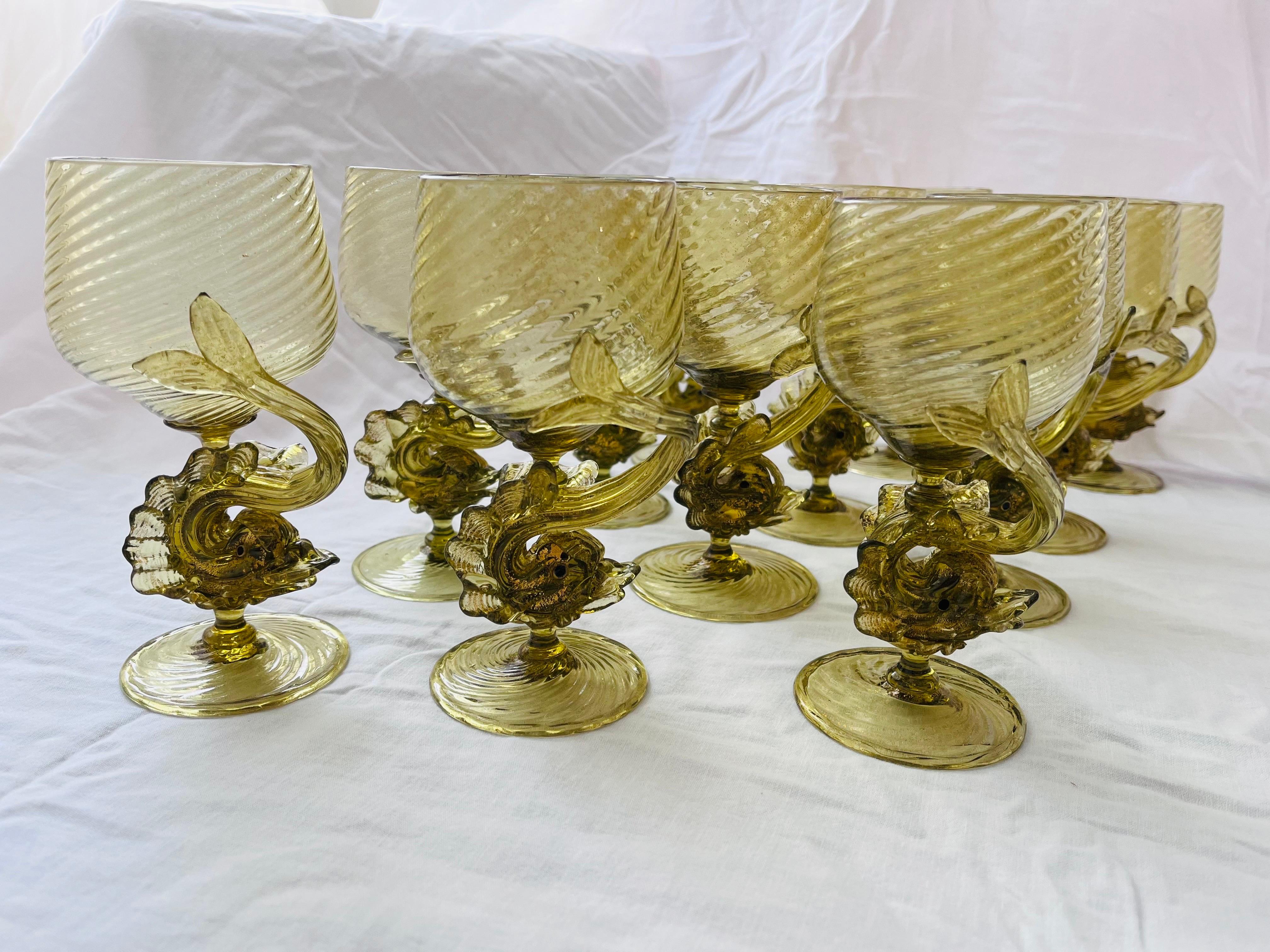 A beautifully ornate set of twelve Antonio Salviati style hand blown glass Venetian Murano style water or wine goblets. The stylized dolphins are the figural stem for each glass. The greenish / amber color is embellished by sparkly gold flecks