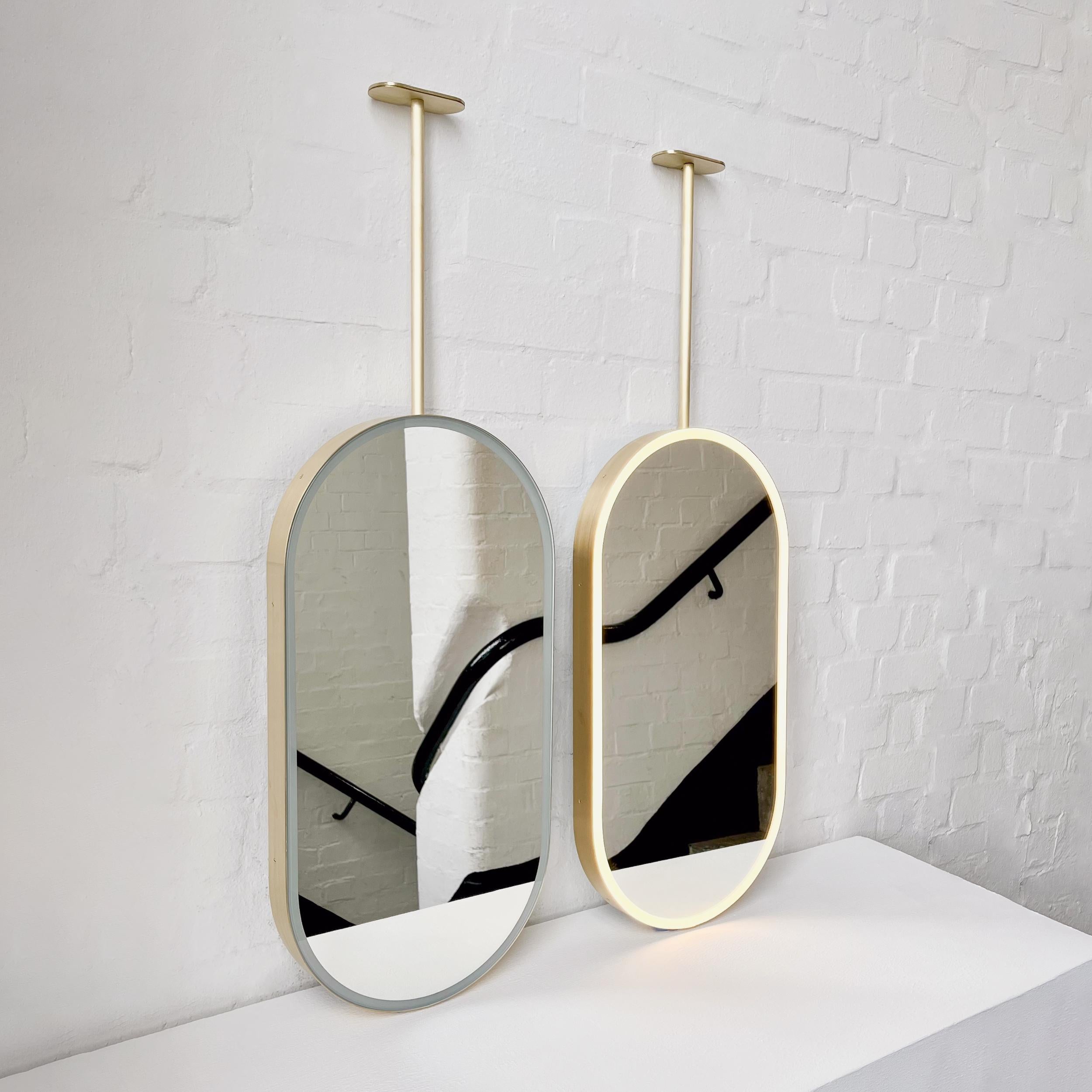 Set of two modern front illuminated Capsula™ ceiling suspended pill shaped mirrors with a minimalist brushed brass frame. Designed and handcrafted in London, UK. This mirror is fully customisable to suit the specific requirements of each