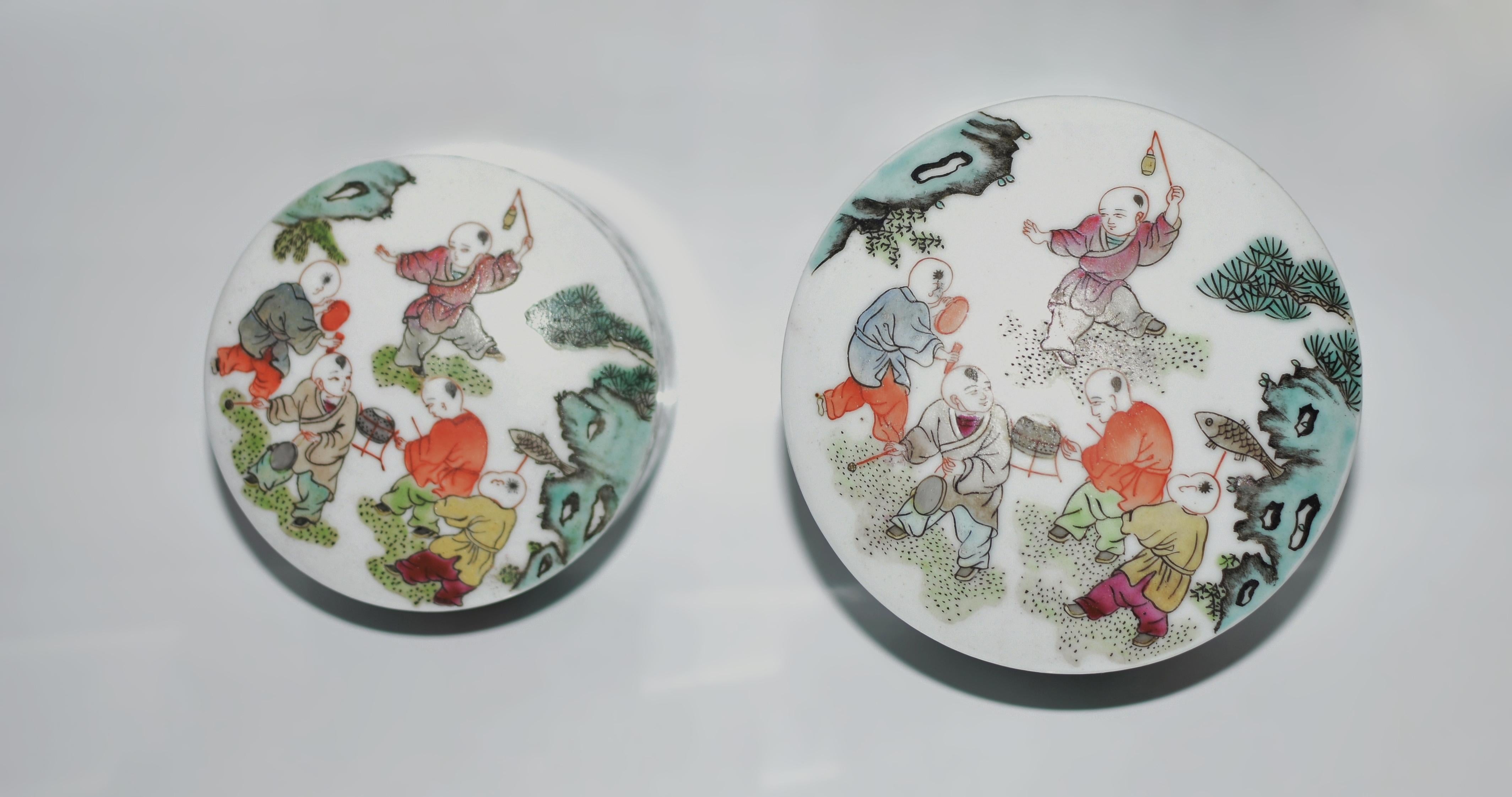 This set of two hand-painted fencai porcelain round boxes from the late Republic era is a charming representation of Chinese artistry and symbolism. Each lid, slightly domed, vividly depicts five boys playing in a garden. One boy holds a fish,