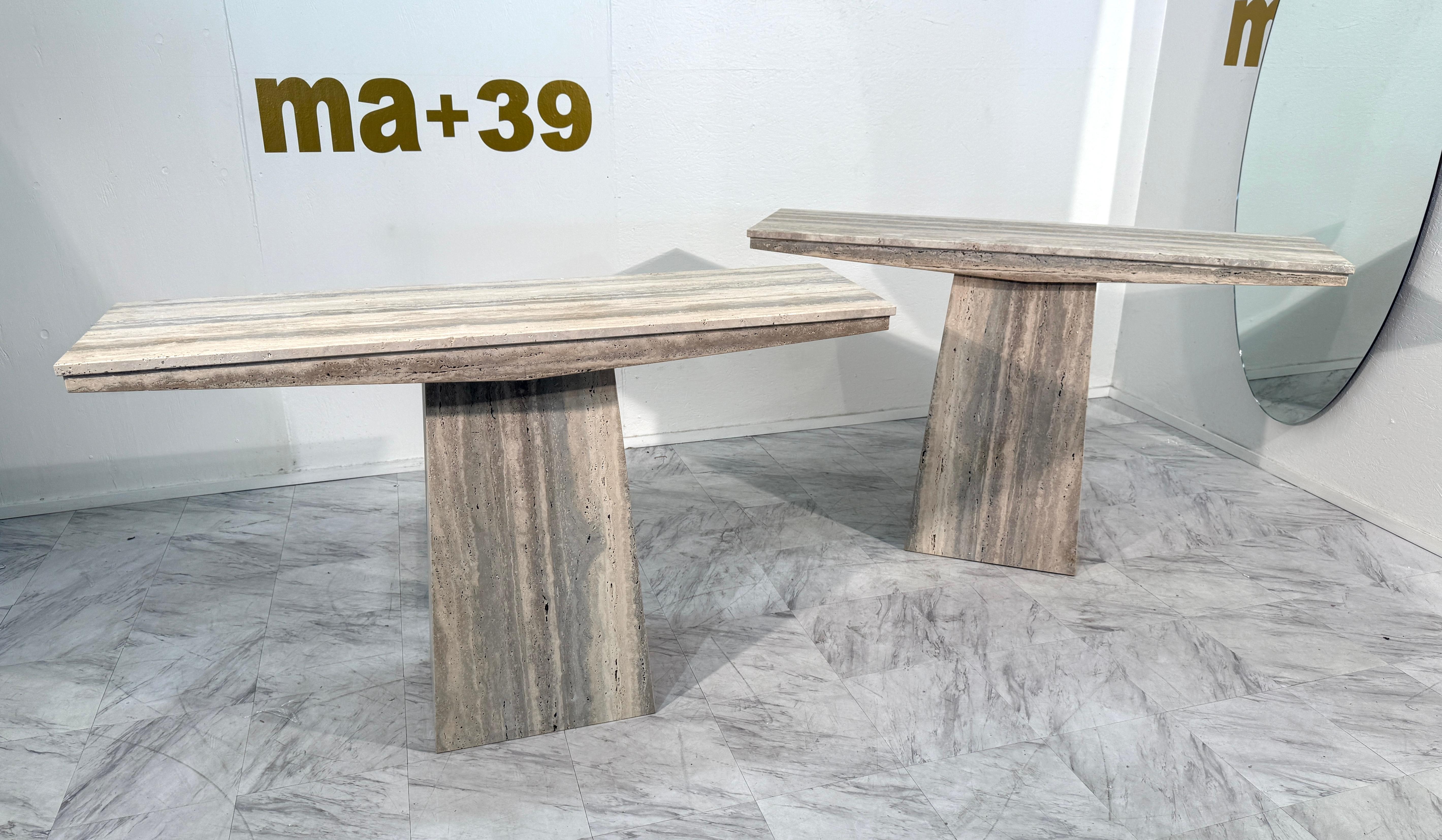 Elegant ensemble de deux tables consoles fabriquées en Italie en pierre naturelle de travertin. Chaque table est dotée d'un plateau rectangulaire aux bords biseautés, reposant sur un solide socle géométrique. Les tons chauds du travertin, beige,