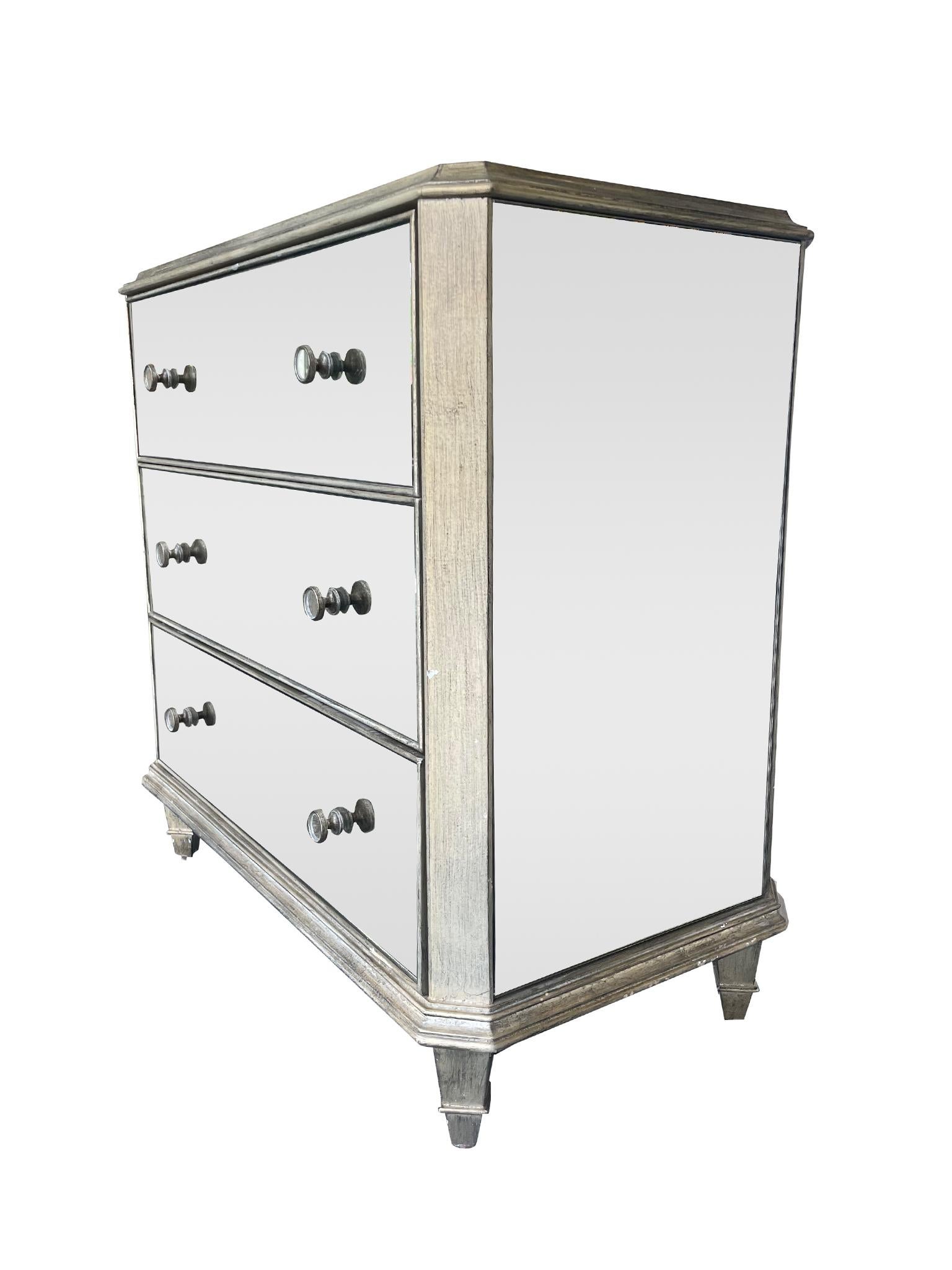 Charming pair of Hollywood Regency style mirrored glass three drawer chests or nightstands. Characterized by their Venetian mirror style panels and silver leaf neoclassical wood base. Designed with low tapered legs, three functional drawers, and