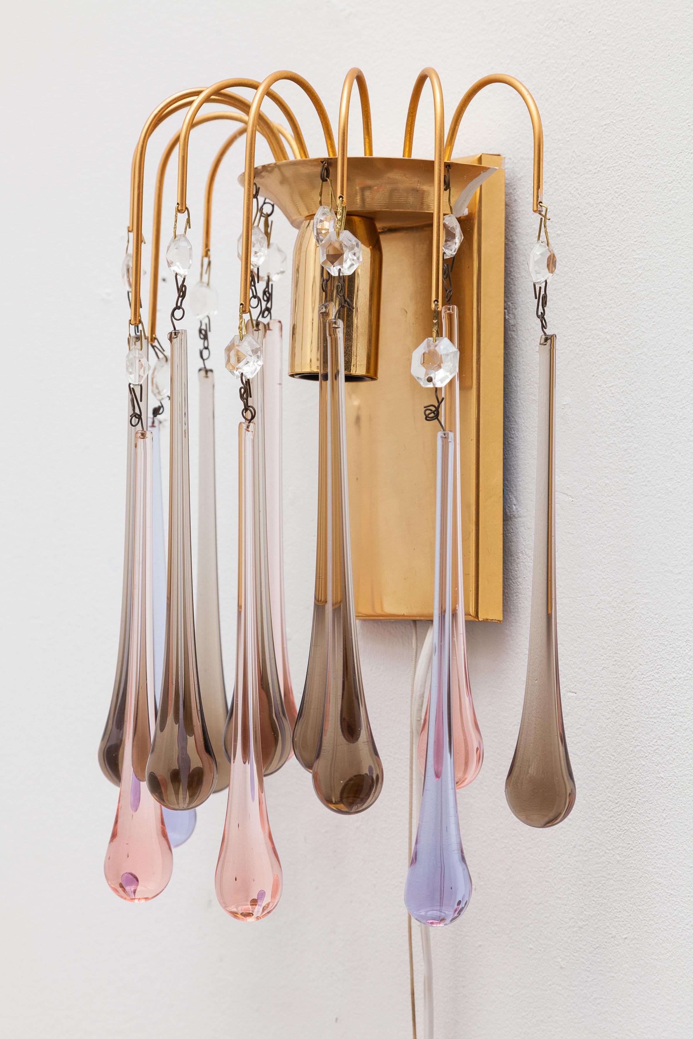 Vintage 1970s wall sconce set. Bright brass frames suspend crystal drops in beautiful shades of lavender, pink and smoked glass.

Both lit by 1 bulb.
Dimension: Sconce:
19W x 30H x 10D cm
Wiring has been checked by a qualified electrician and